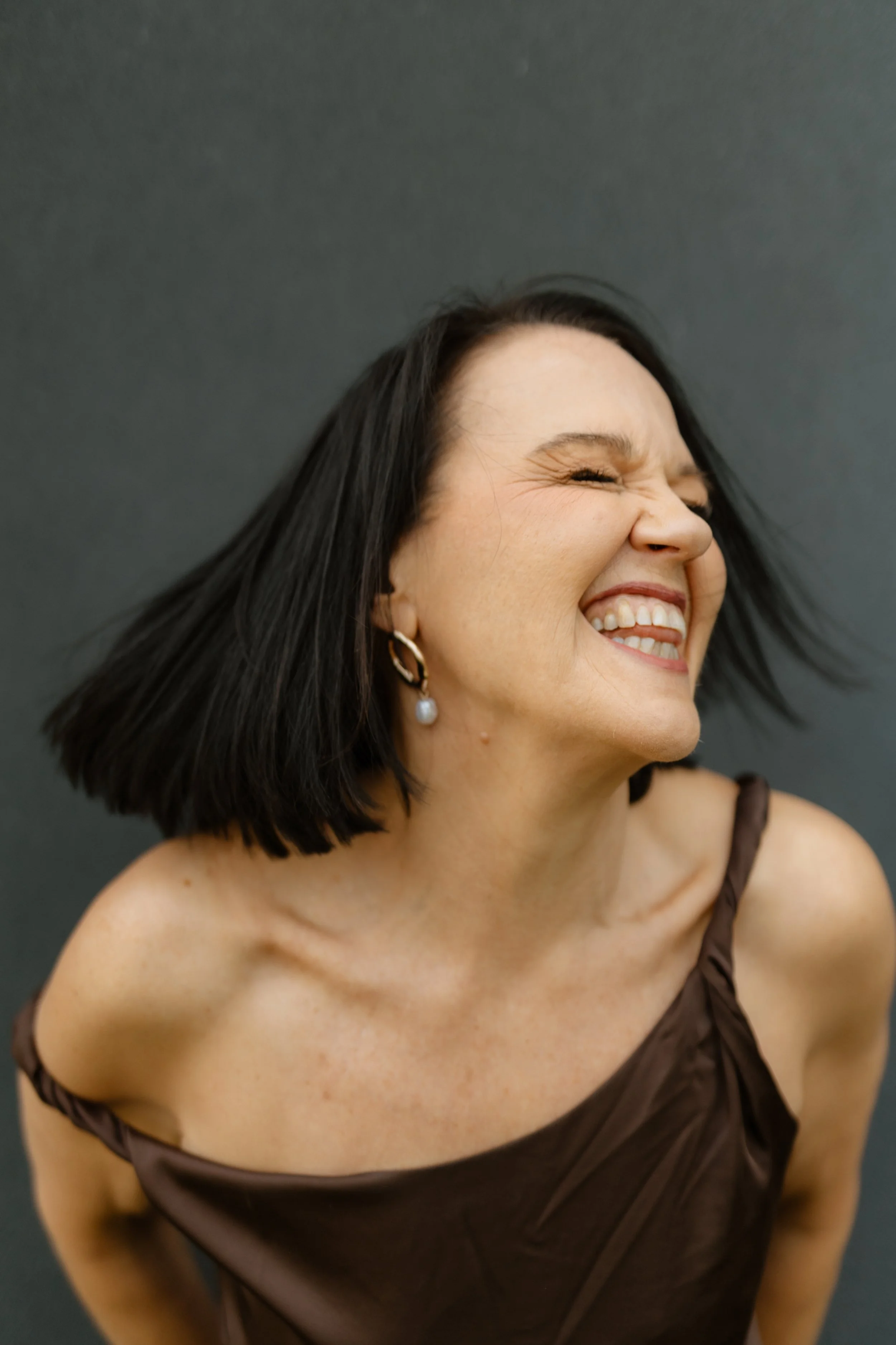 A woman smiling broadly with her eyes closed and head turned to the side, wearing a dark brown dress and pearl earrings, against a plain dark background.