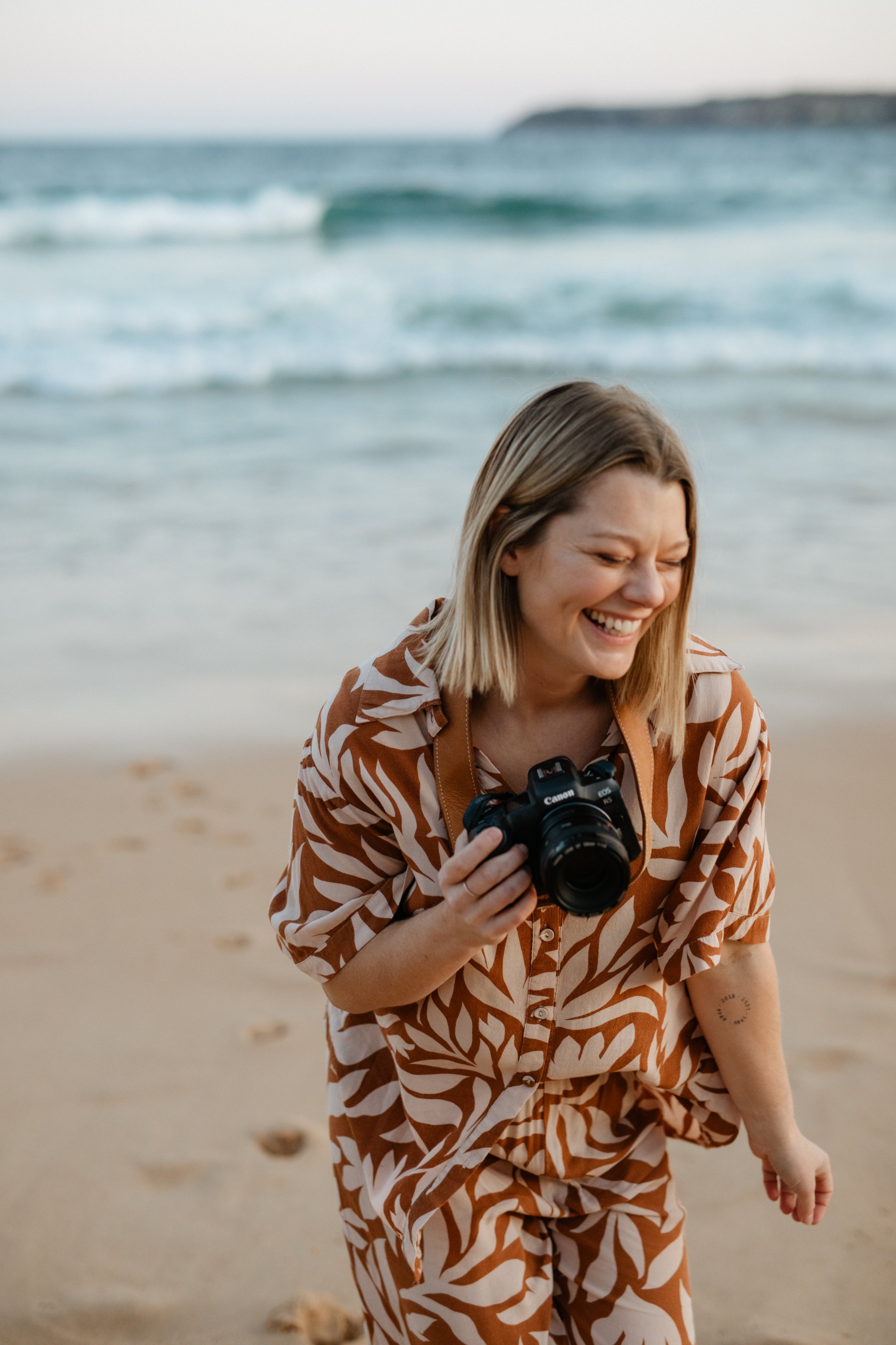 A woman with blonde hair, smiling while holding a camera, standing on a sandy beach with waves and a distant landform in the background.