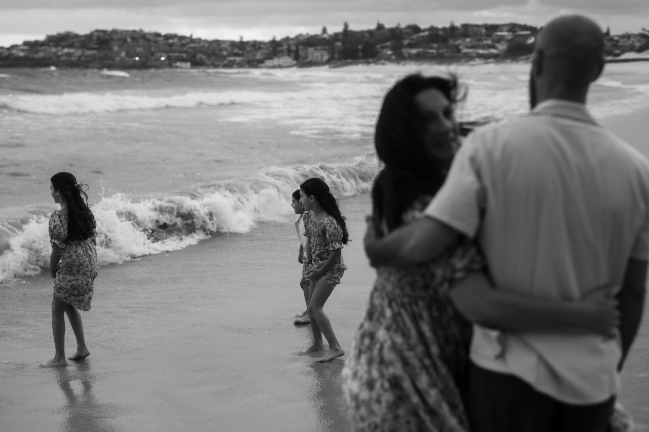 A black-and-white photo of a beach scene with a couple hugging in the foreground and three women standing at the water's edge with waves in the background.