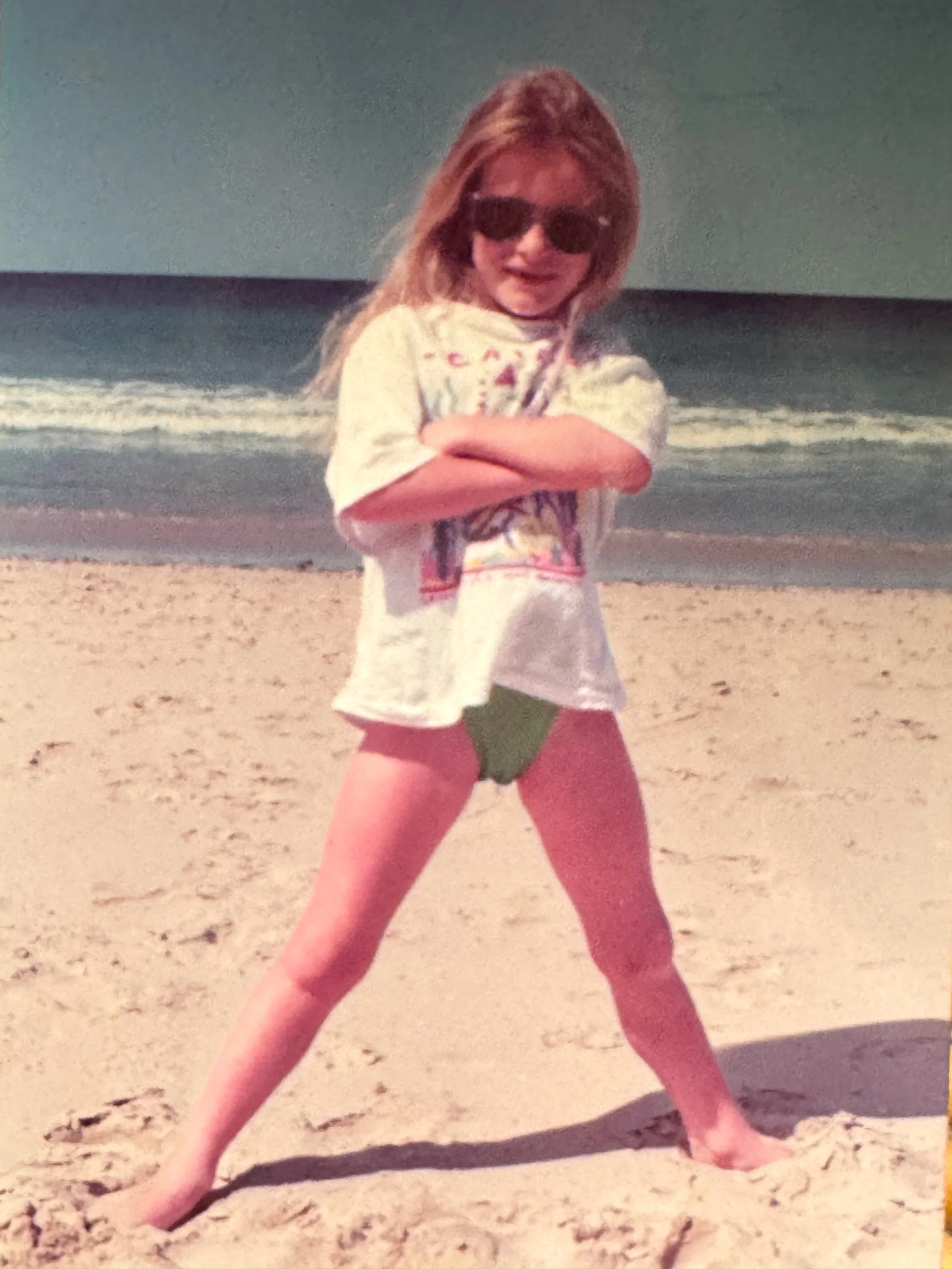 A young girl standing on a sandy beach with arms crossed wearing sunglasses, a colorful t-shirt, and green bikini bottoms.