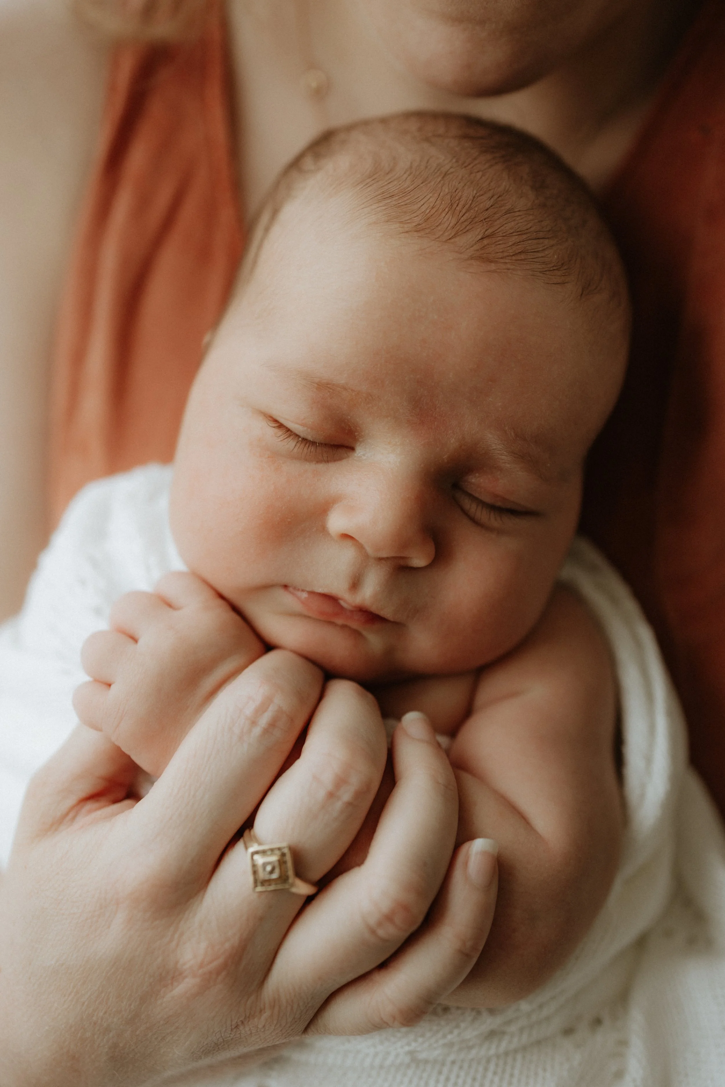 Close-up of a sleeping baby with a hand gently holding the baby's hand, wearing a gold ring with a square design.