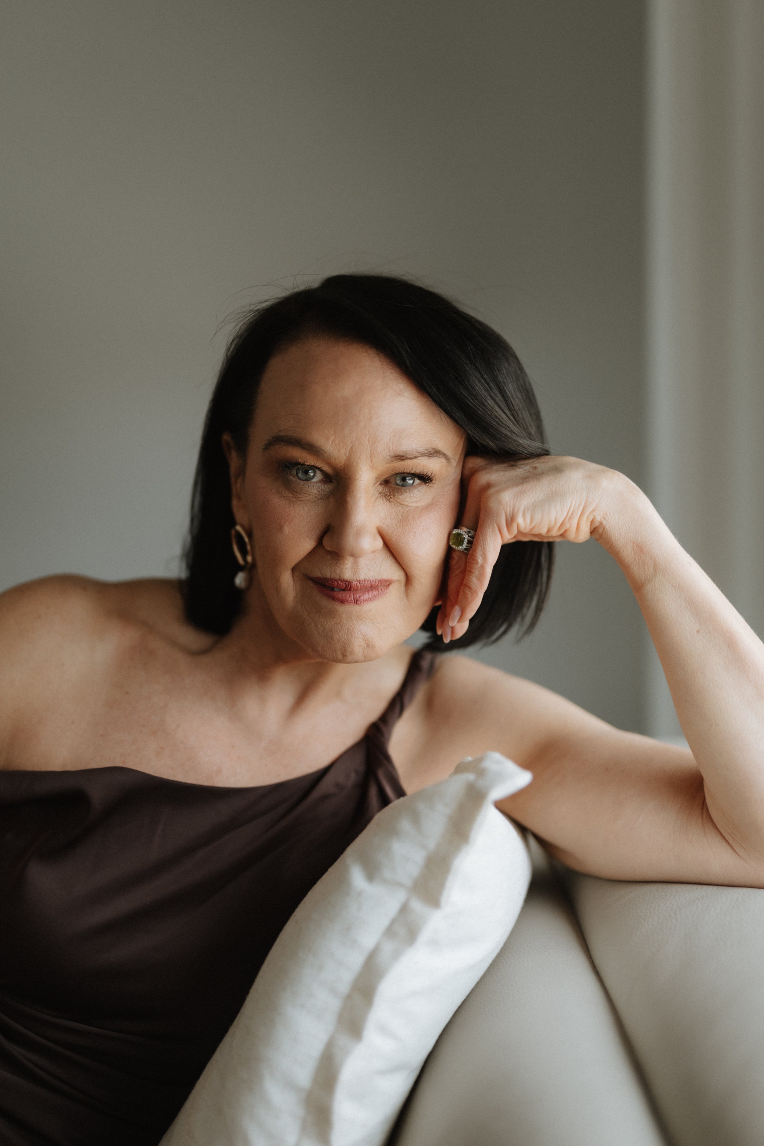 A woman with dark hair and blue eyes sitting on a beige couch, resting her head on her hand, wearing a dark satin sleeveless top, and gold hoop earrings.
