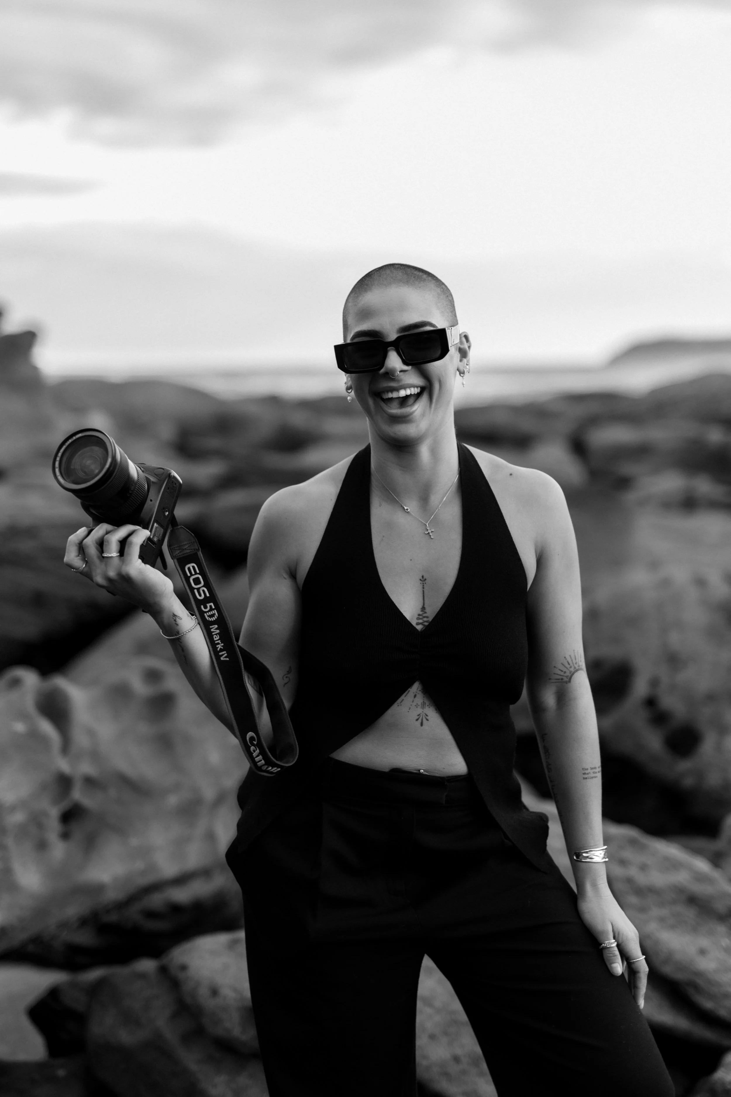 A smiling woman with a shaved head wearing large sunglasses, a black halter top, and multiple jewelry pieces, holding a Canon camera strap, standing outdoors in a rocky landscape with hills visible in the background.