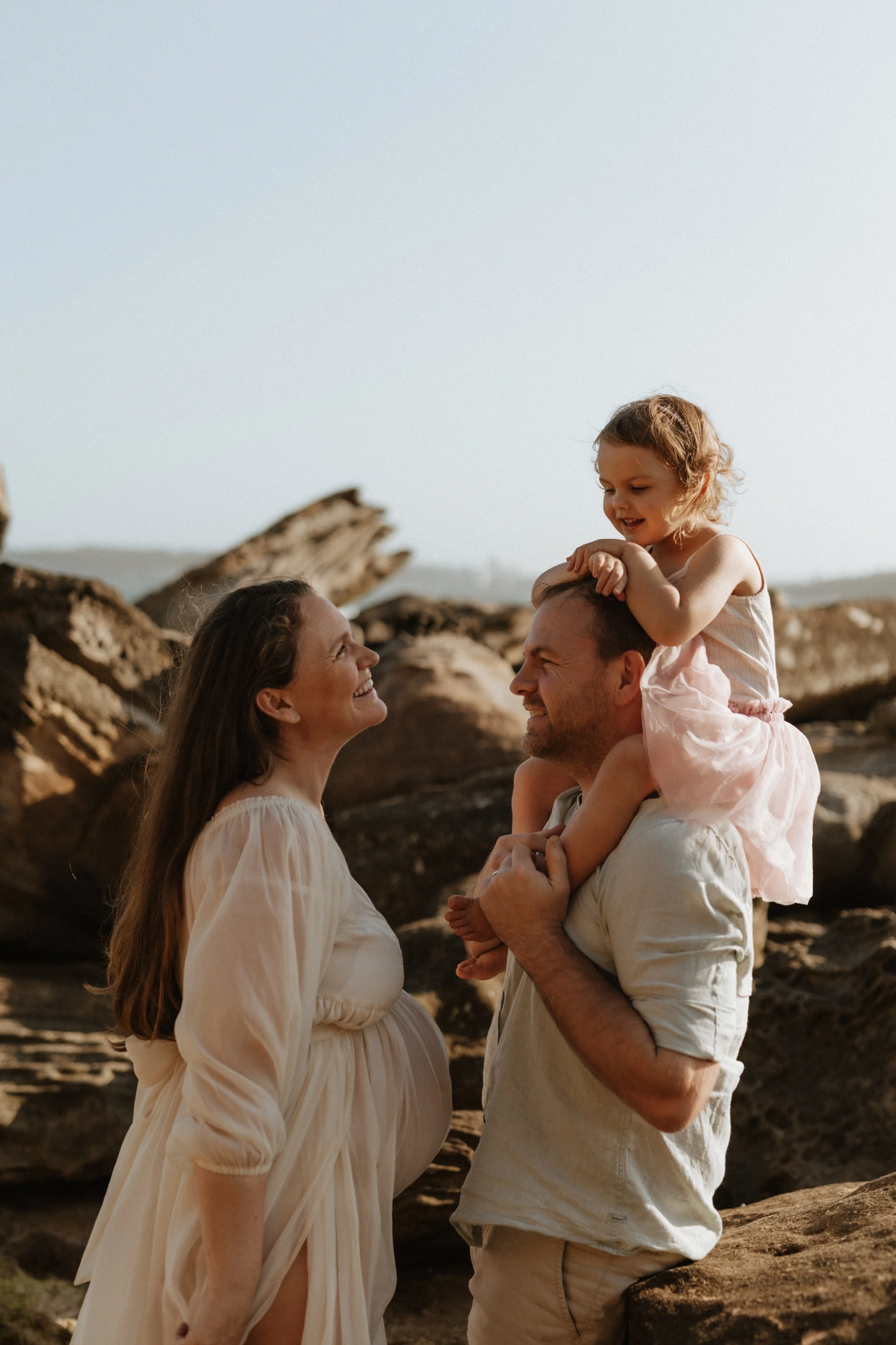 A family of four at the beach, with large rocks in the background. The mother, father, and daughter are smiling and looking at each other. The daughter is sitting on the father's shoulders.