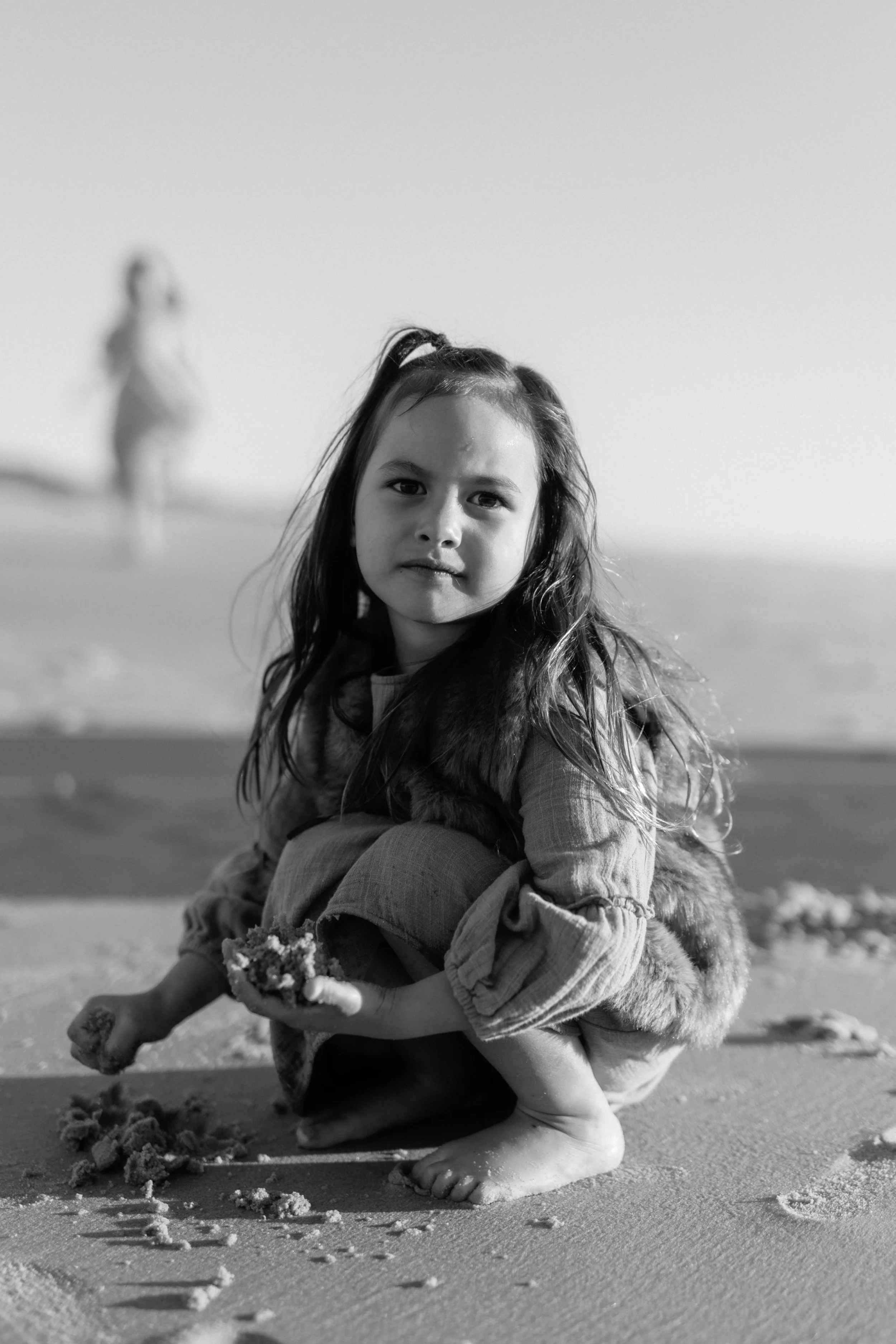 A young girl with long hair in a ponytail, crouching on a sandy beach, holding a handful of sand, with more sand scattered around her. She is wearing a jacket and appears to be playing with the sand. In the background, there is a blurred figure of another person walking along the beach.