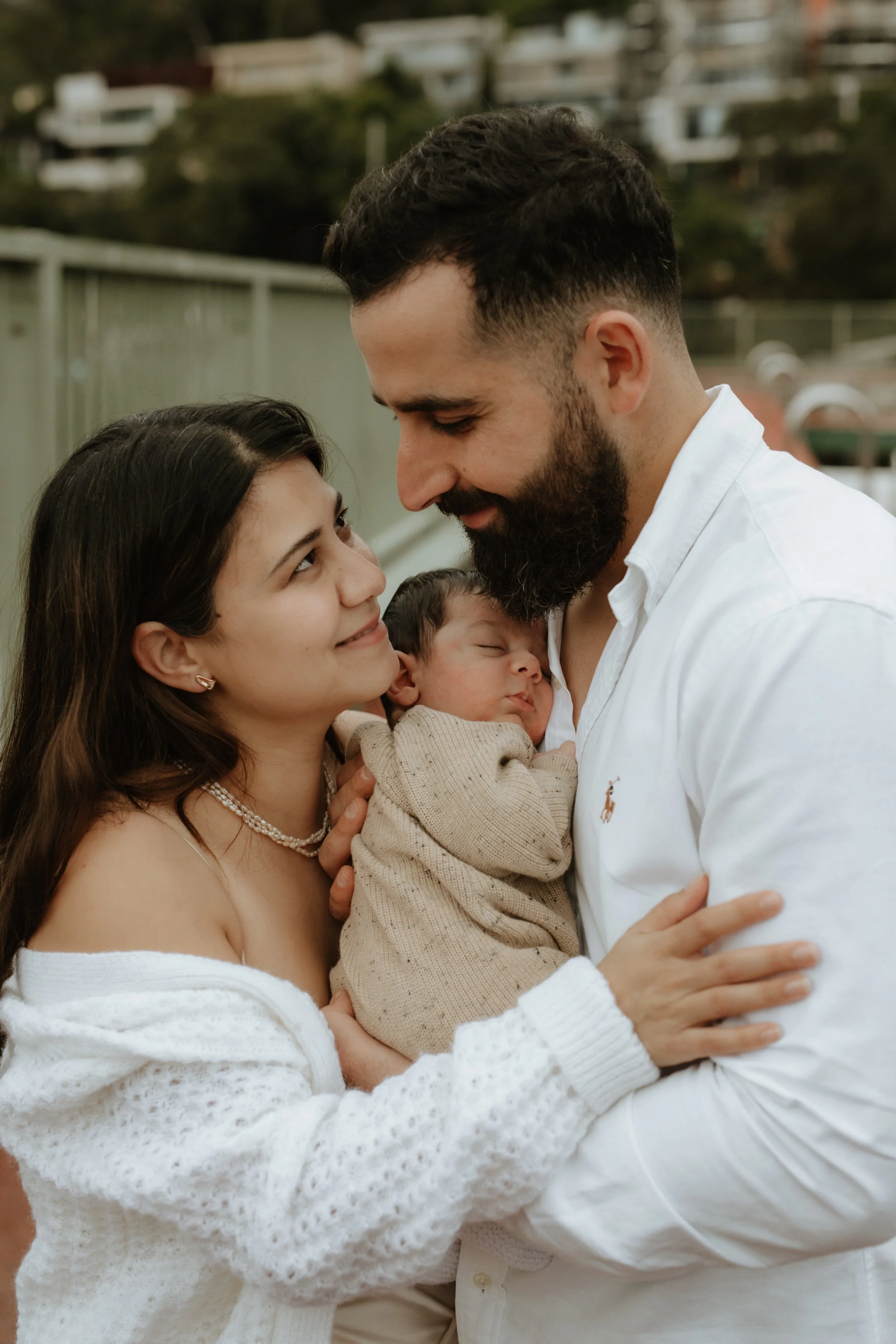 A woman and a man holding a sleeping baby, looking into each other's eyes, outdoors.