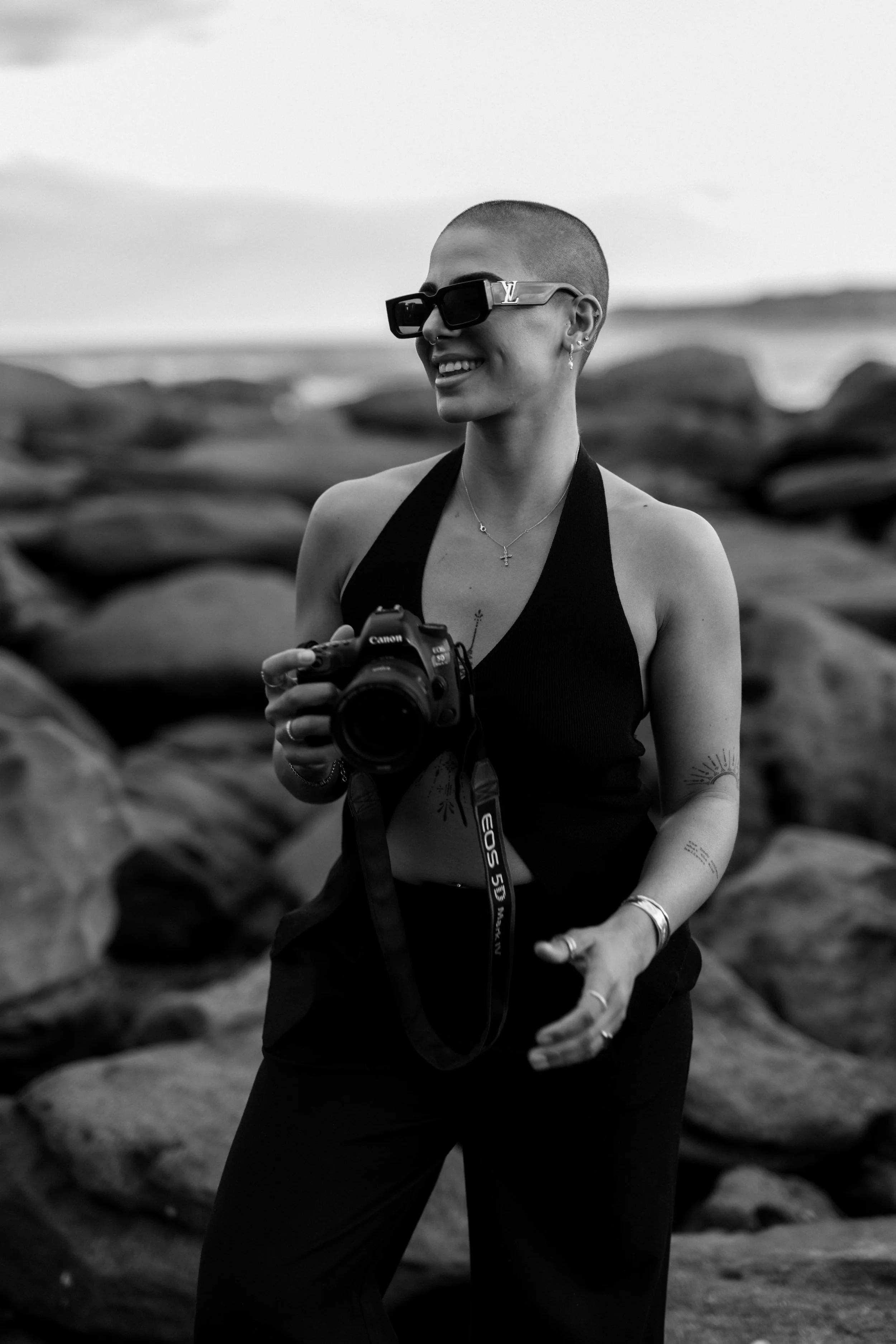 A woman with a shaved head wearing large dark sunglasses, smiling and holding a camera, outdoors with rocks in the background.