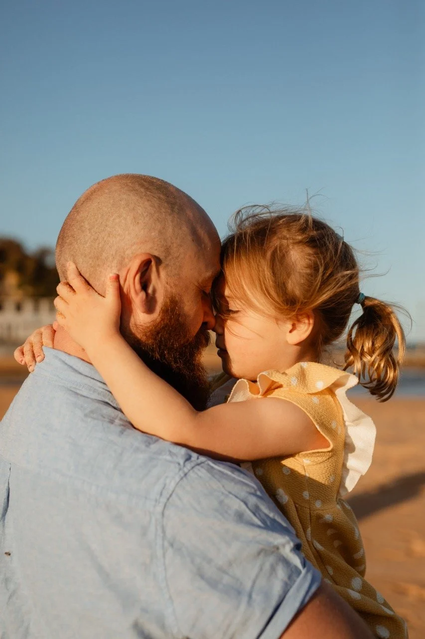 A man with a beard holding a young girl with pigtails close, with their foreheads touching outdoors on a sandy area under a clear blue sky.