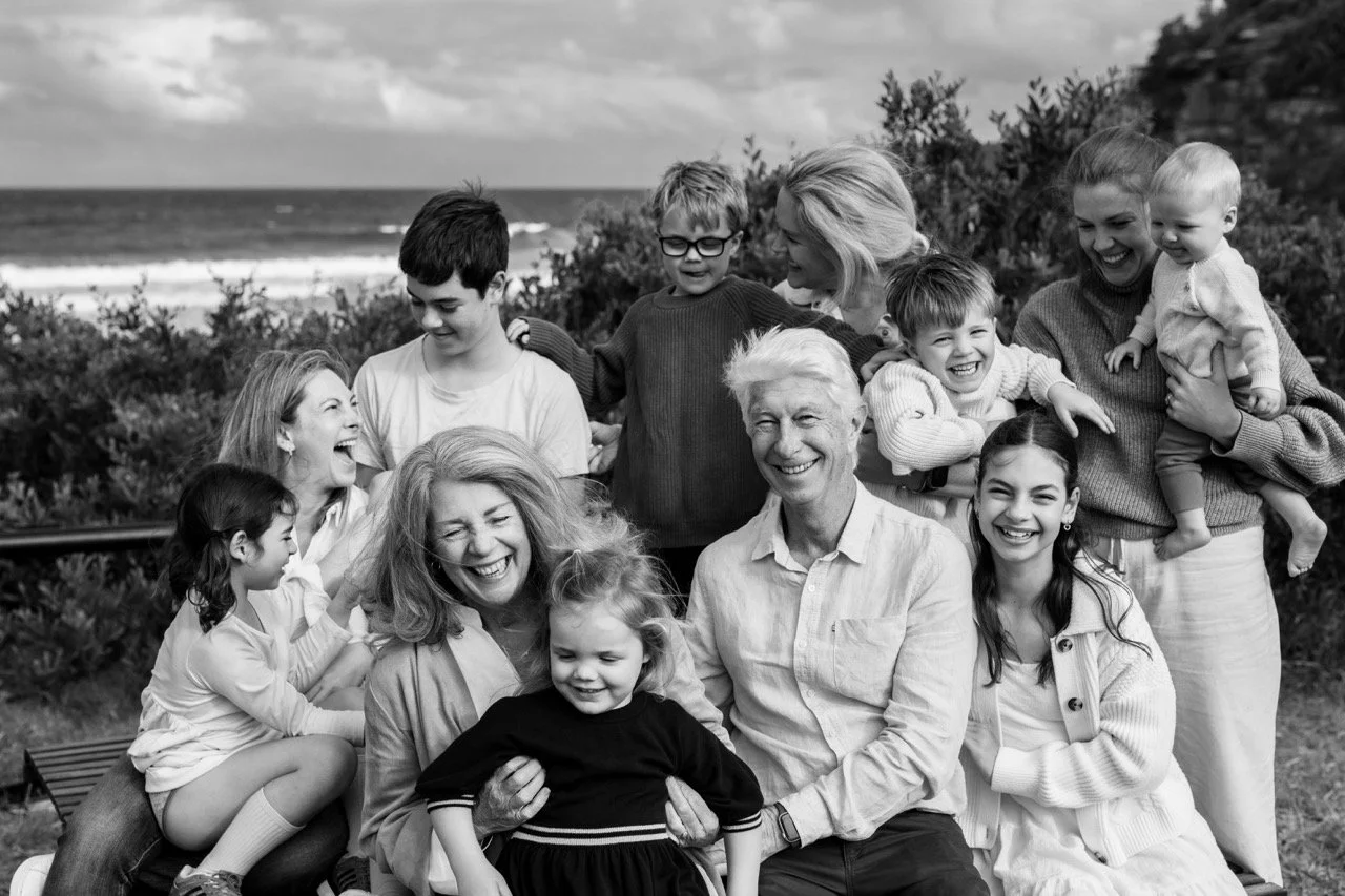 A black and white photo of a large family enjoying time together outdoors near the beach, with parents, children, and grandchildren smiling and interacting.