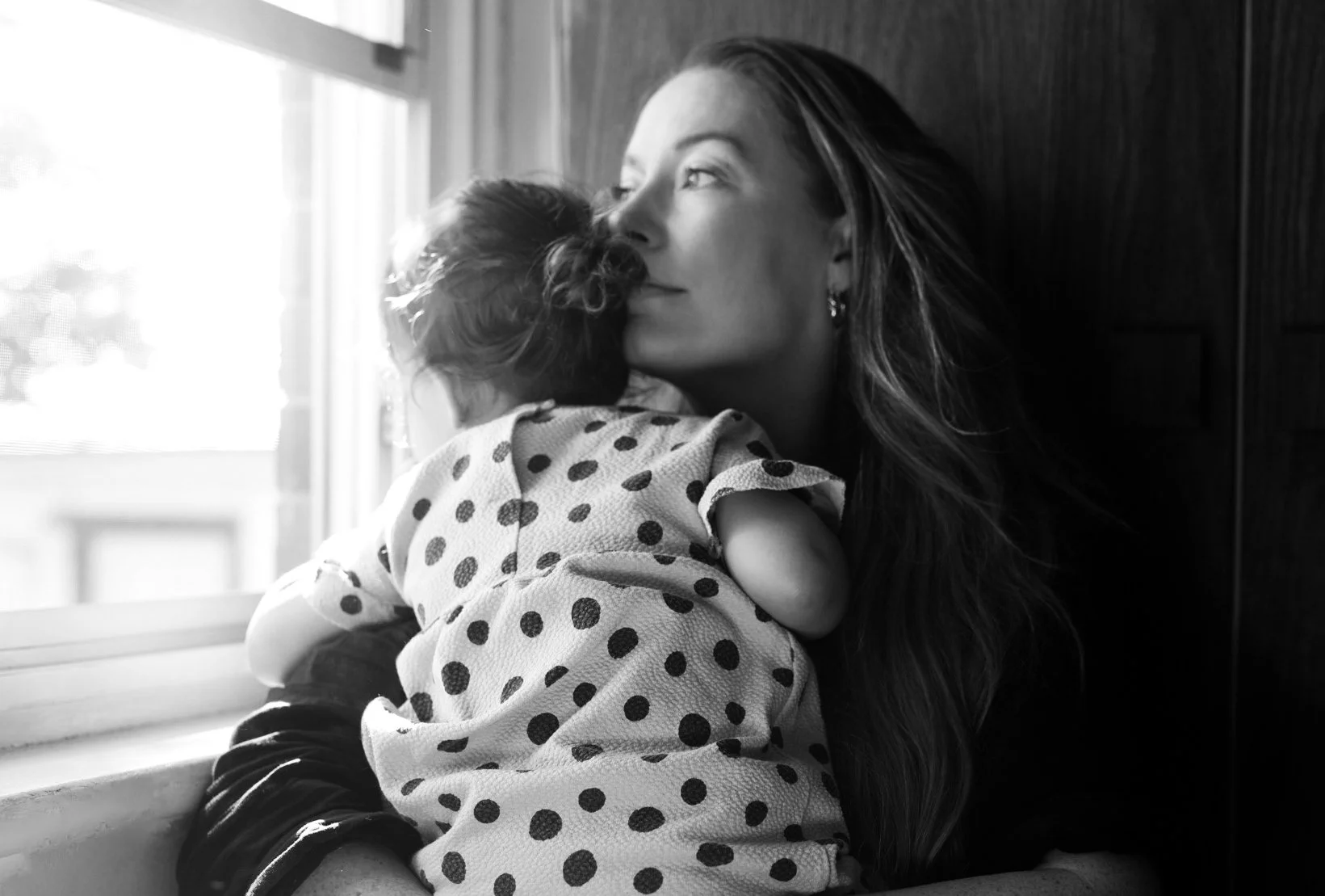 A woman with long hair holding a young child close to her face, both sitting near a window. The image is in black and white.