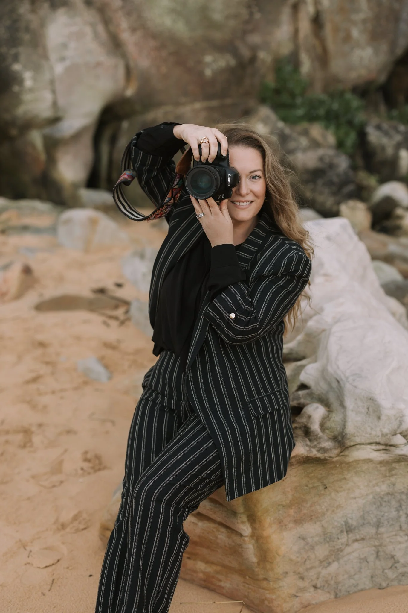 A woman with curly hair, wearing a black and white pinstripe suit, is smiling and holding a camera to her eye, taking a picture outdoors near large rocks.