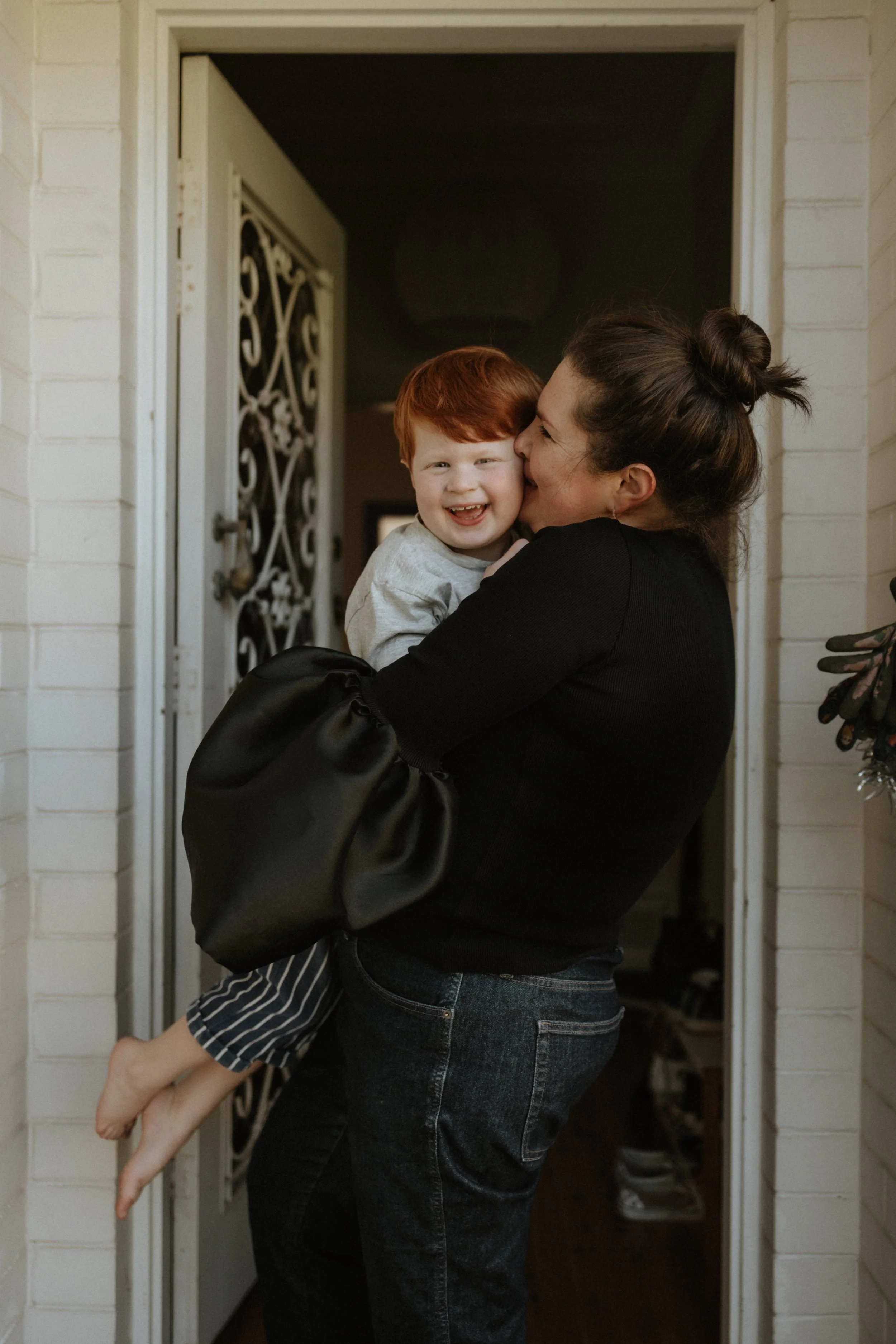 A woman is holding a young boy in her arms, kissing his cheek, by a doorway. The boy is smiling and wearing striped pajama pants. The woman has her hair in a bun and is wearing a black top and jeans.