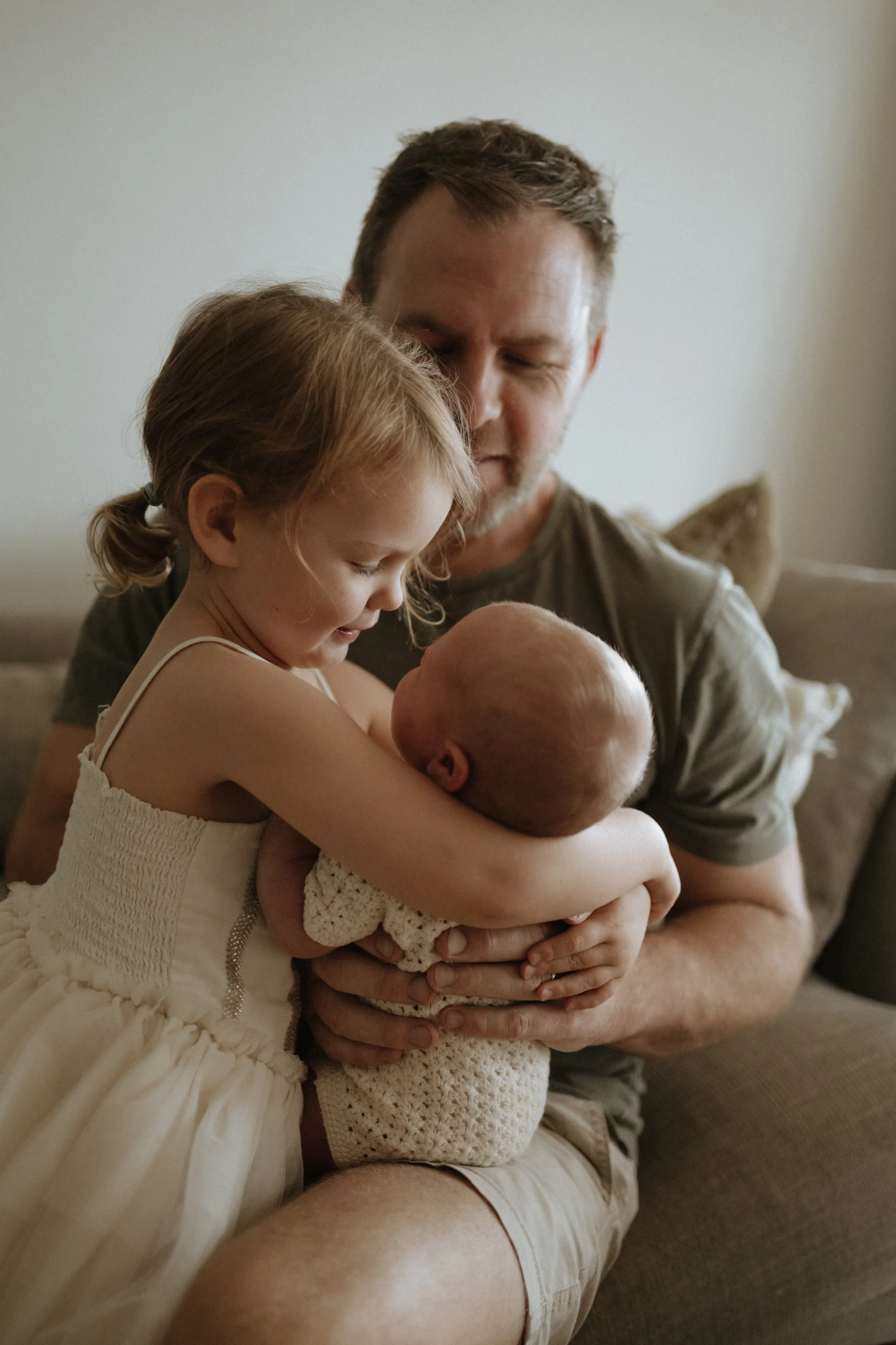 A man holding a sleeping newborn baby in his arms while a young girl, possibly an older sibling, leans in to look at the baby. The scene is warm and tender, set in a cozy indoor environment.