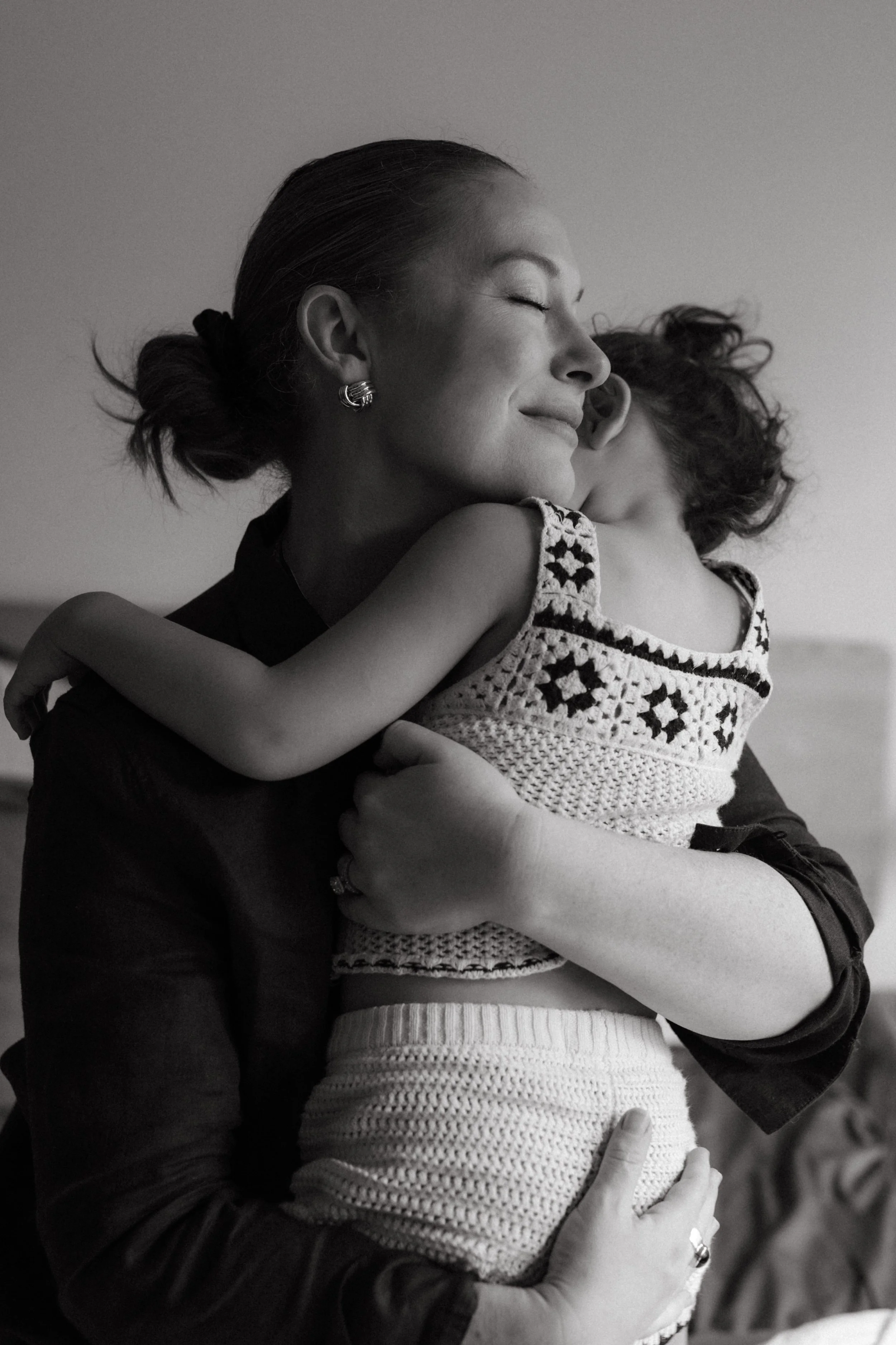 A woman with her eyes closed hugging a child who is wearing a sleeveless dress. The woman has her hair tied back and is wearing earrings, and the child has curly hair.