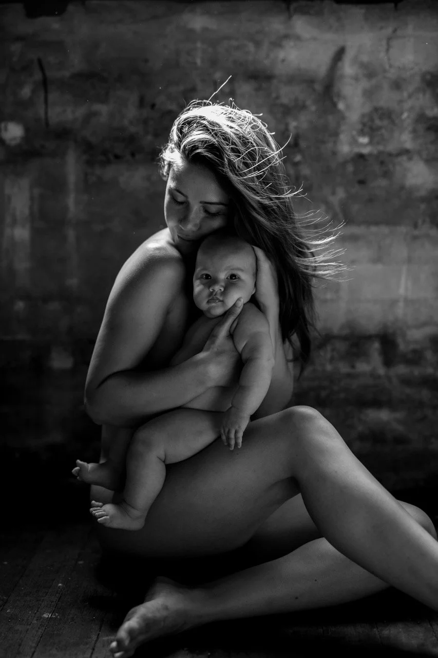 A black-and-white photo of a woman holding a baby, both sitting on the floor with a wooden background. The woman is sitting with her legs crossed, holding the baby close to her chest.