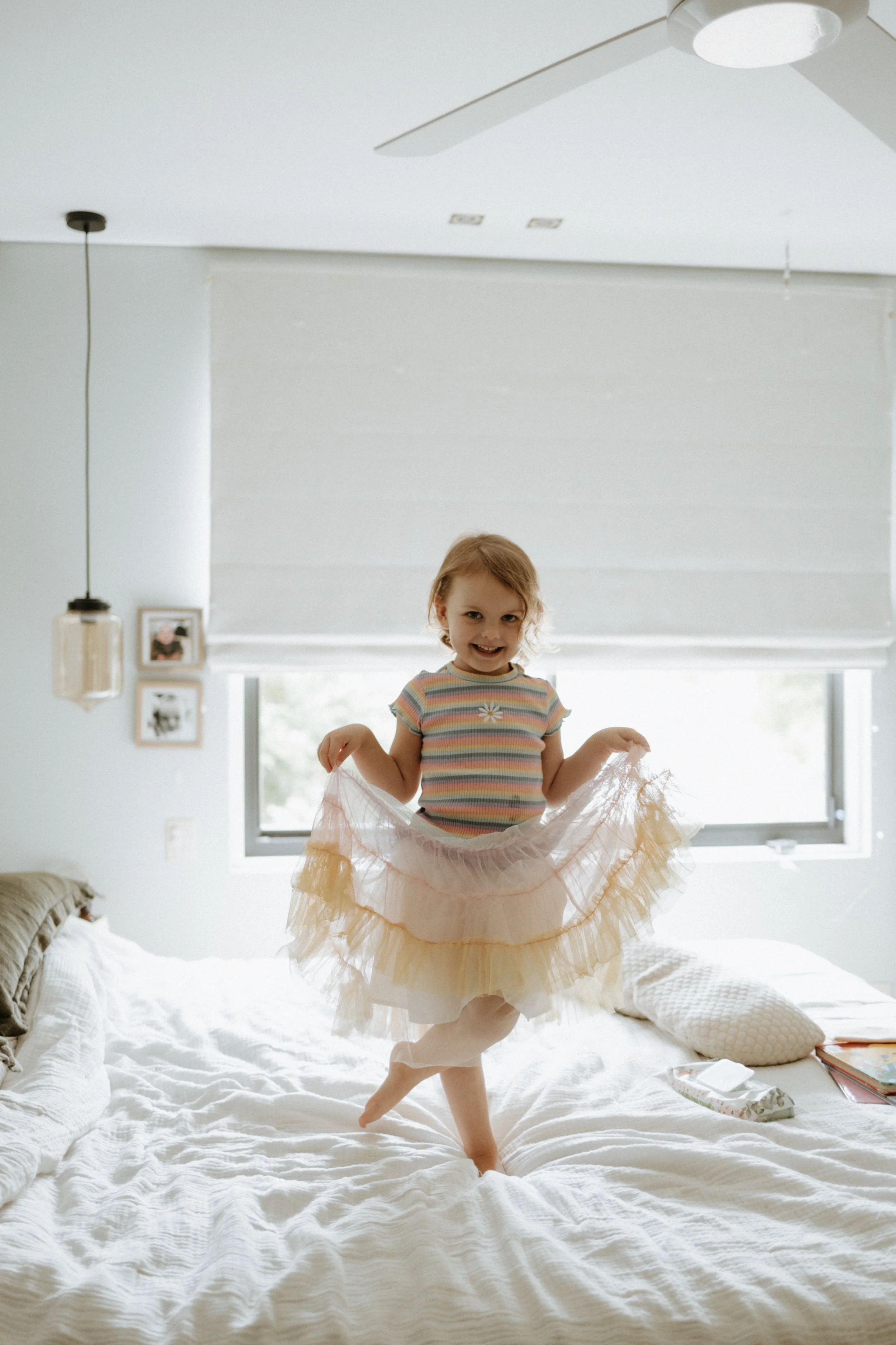 A young girl with red hair wearing a striped shirt and a frilly beige tutu is standing on a white bed, smiling at the camera, in a bright bedroom with a window and framed pictures on the wall.