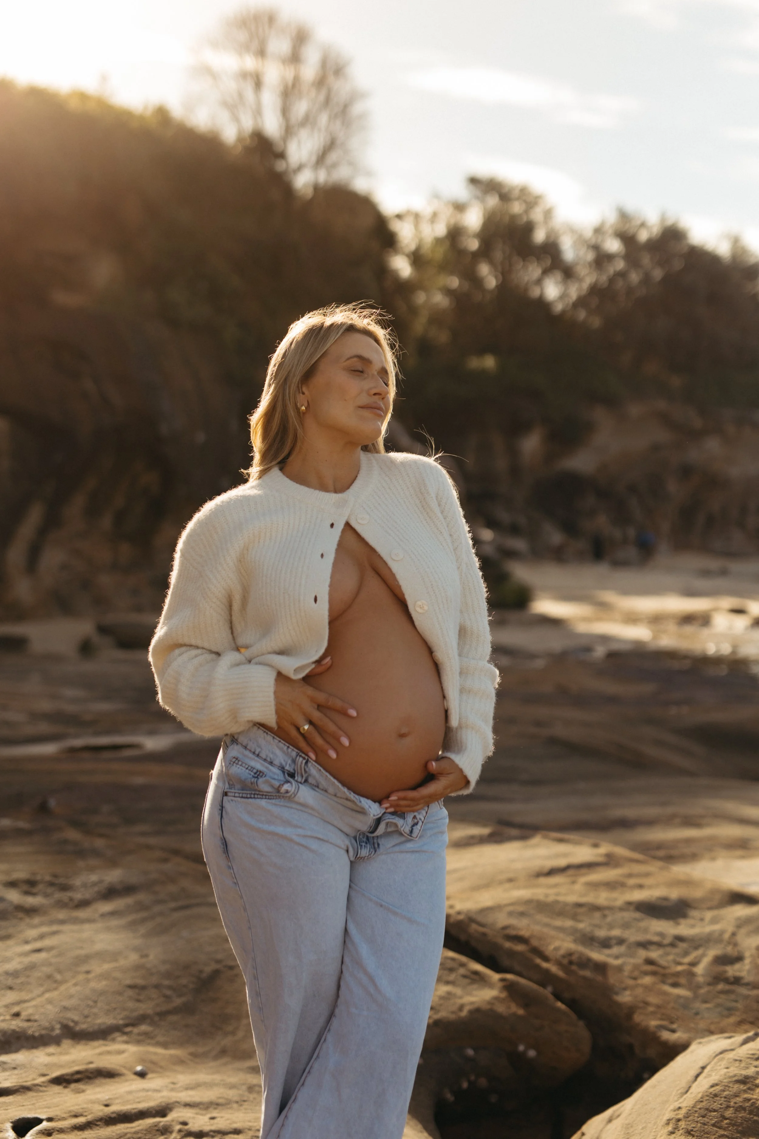 A woman with blonde hair standing outdoors on a rocky beach during sunset, holding her pregnant belly with both hands, wearing a cream-colored cardigan and light blue jeans, with her eyes closed and a peaceful expression.