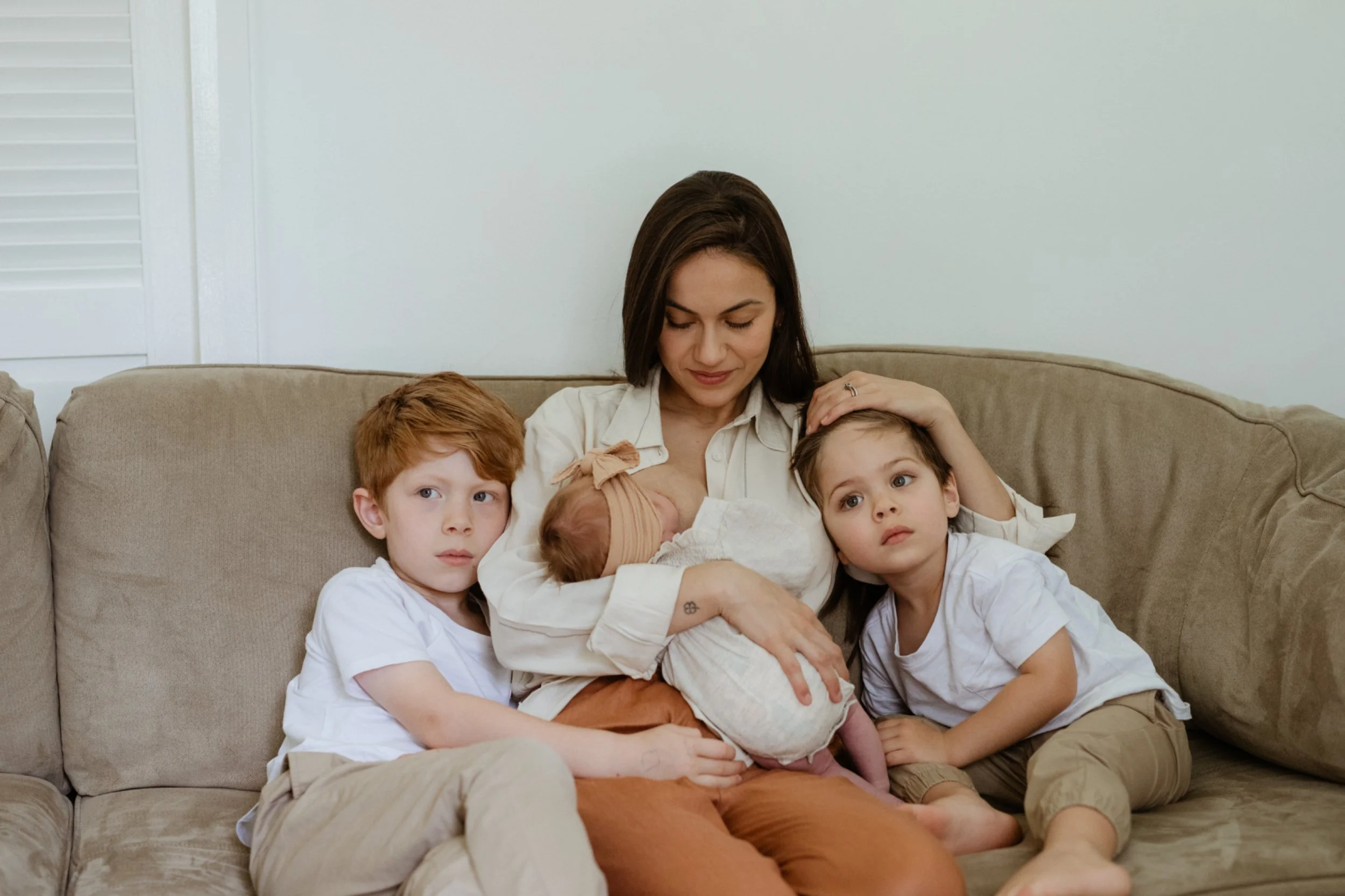 A woman with dark hair holding a newborn baby and sitting on a beige sofa, surrounded by three young children with light brown hair. All are dressed in white shirts and seated together indoors against a plain wall.