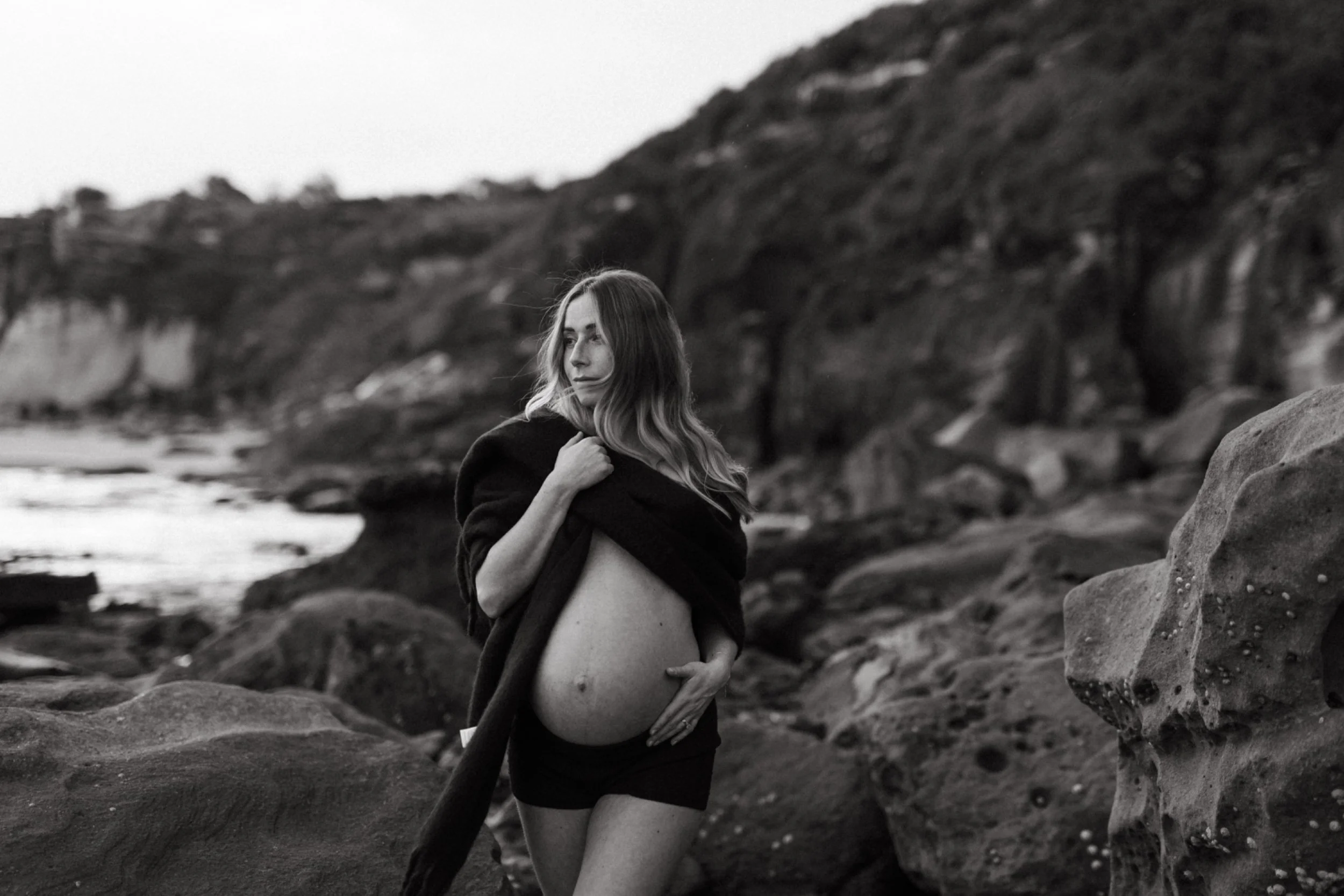 A woman standing on rocks near the ocean, holding her shirt open to expose her pregnant belly, with a rocky landscape in the background in black and white.