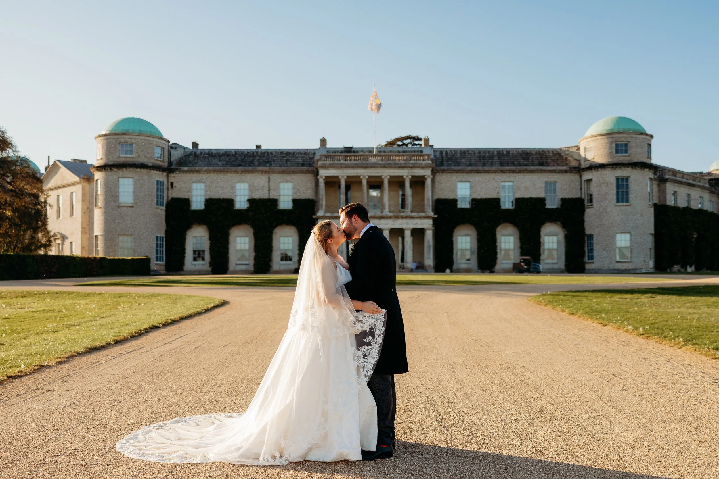 A bride and groom are kissing in front of a historic building with green domed roofs and ivy-covered walls, on a sunny day at Goodwood Estate, Sussex.