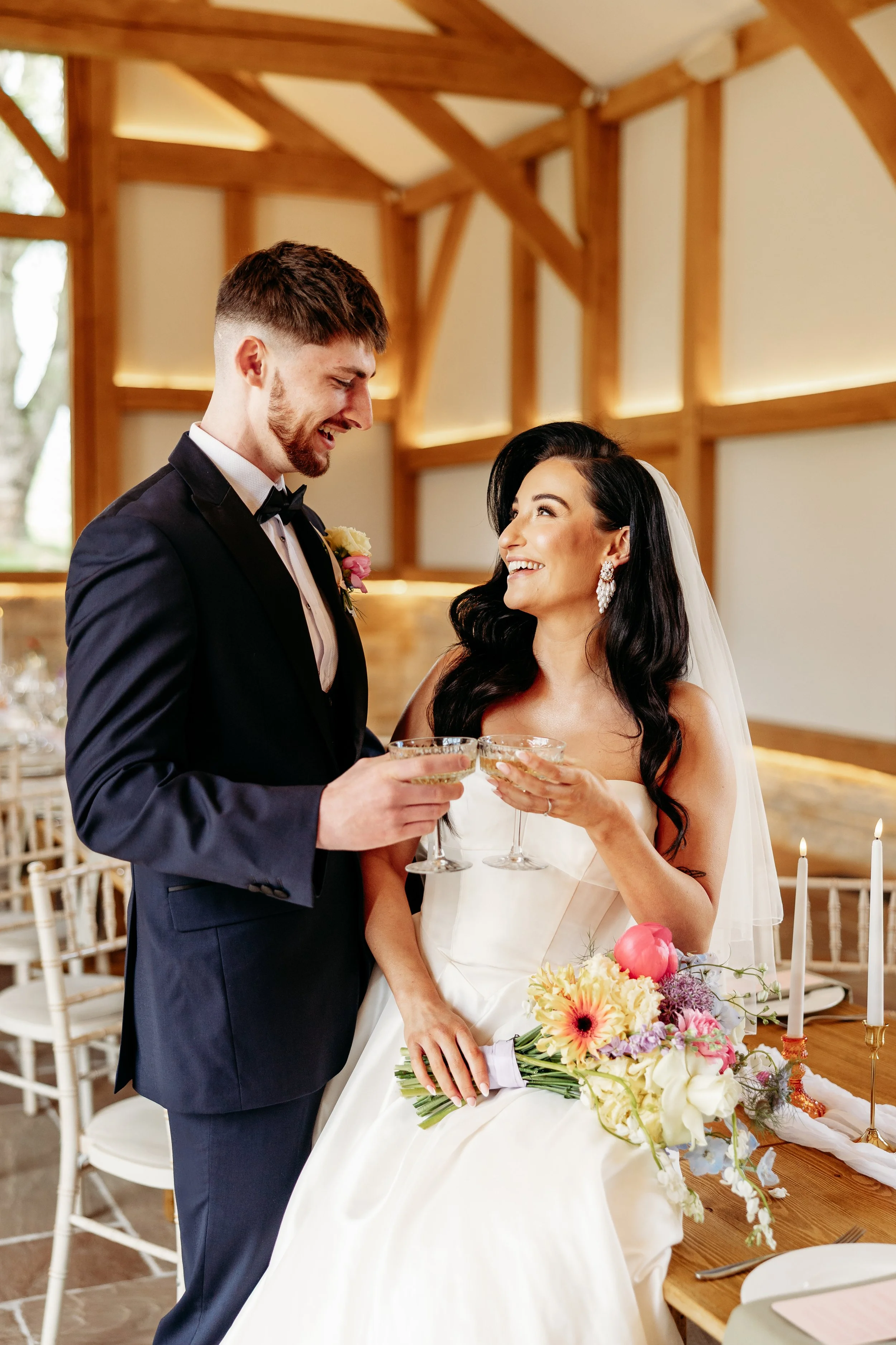 A bride and groom smiling and holding champagne glasses at their wedding reception inside a rustic venue with wooden beams. The bride is seated with a bouquet of flowers on her lap, and the groom stands beside her.