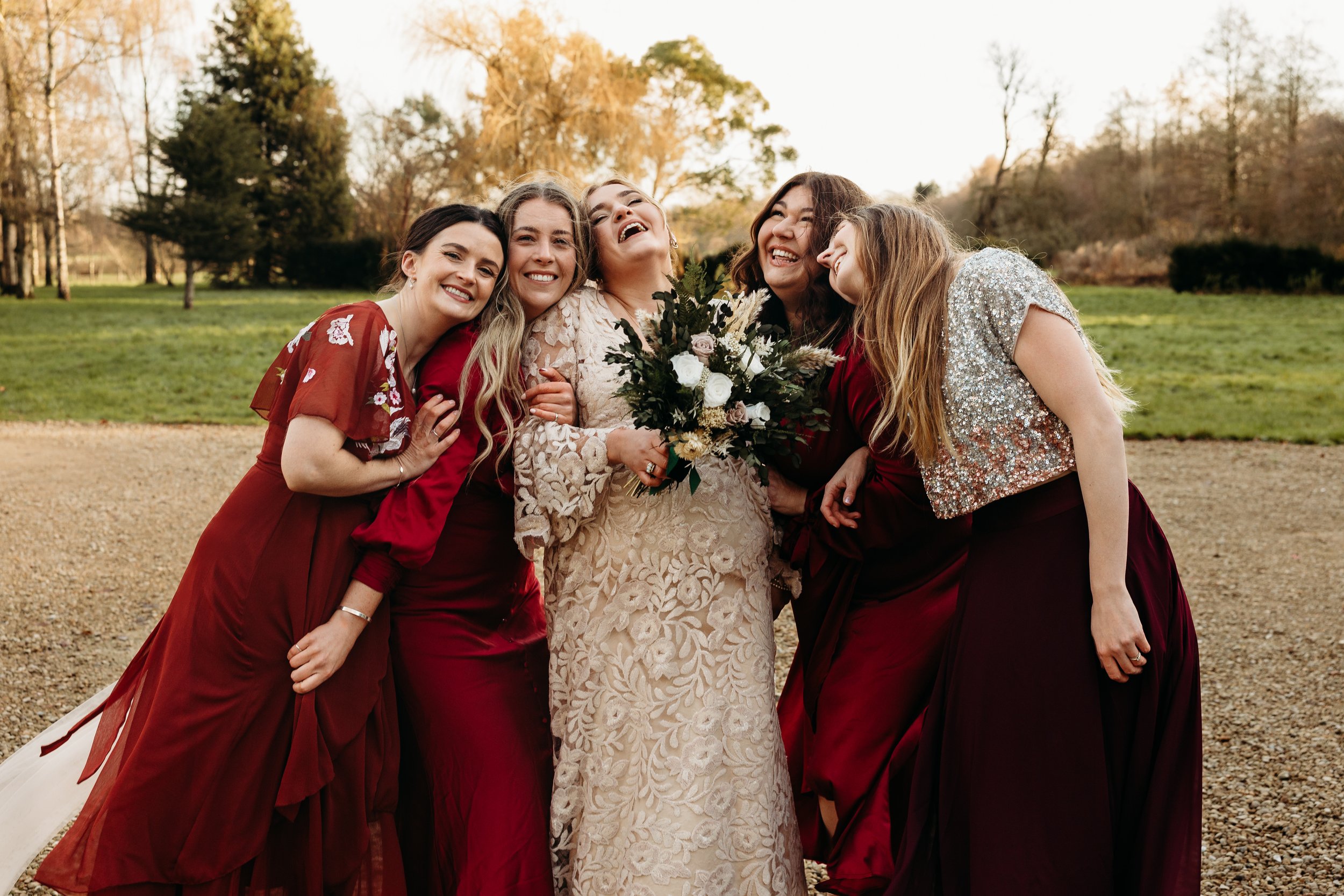 A bride and five bridesmaids smiling and hugging outdoors in a park with trees in the background, during sunset or late afternoon.