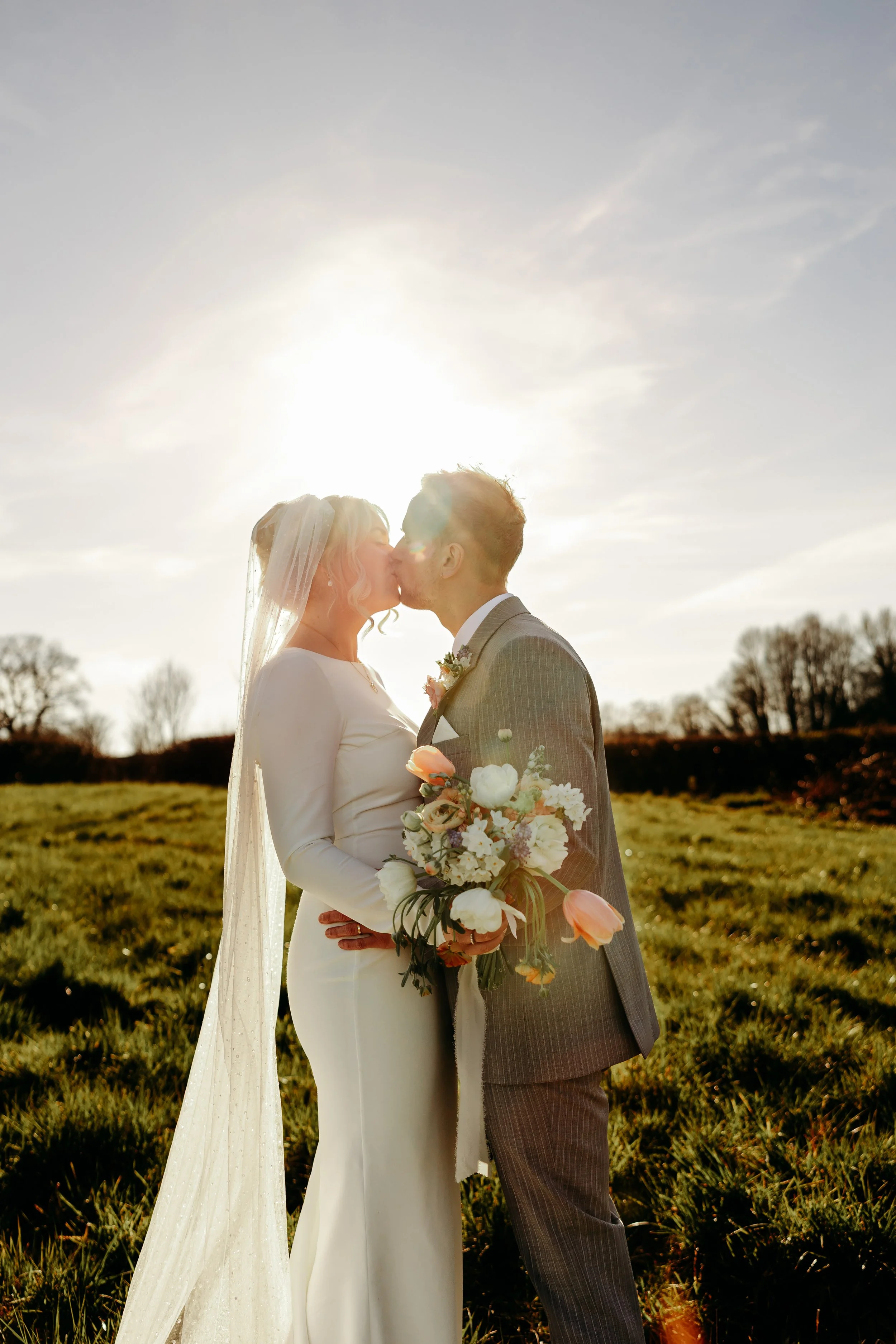 A bride and groom kiss in a sunlit outdoor setting, with the bride holding a bouquet of flowers and wearing a white dress with a veil, and the groom in a gray suit.