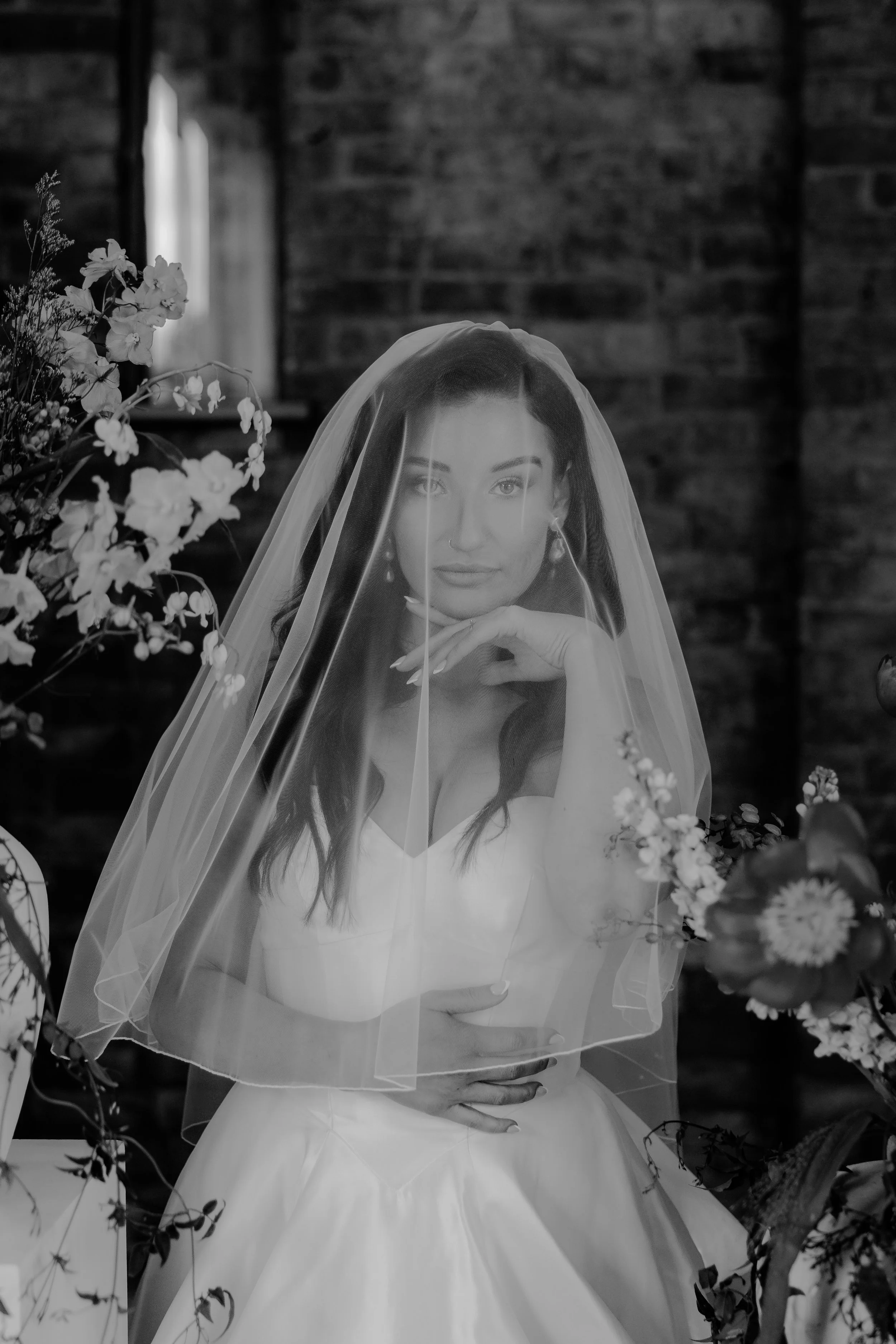 A woman wearing a wedding dress and veil, sitting among flowers with a brick wall in the background at Mickleton Hill Farm, Cheltenham, Cotswolds.