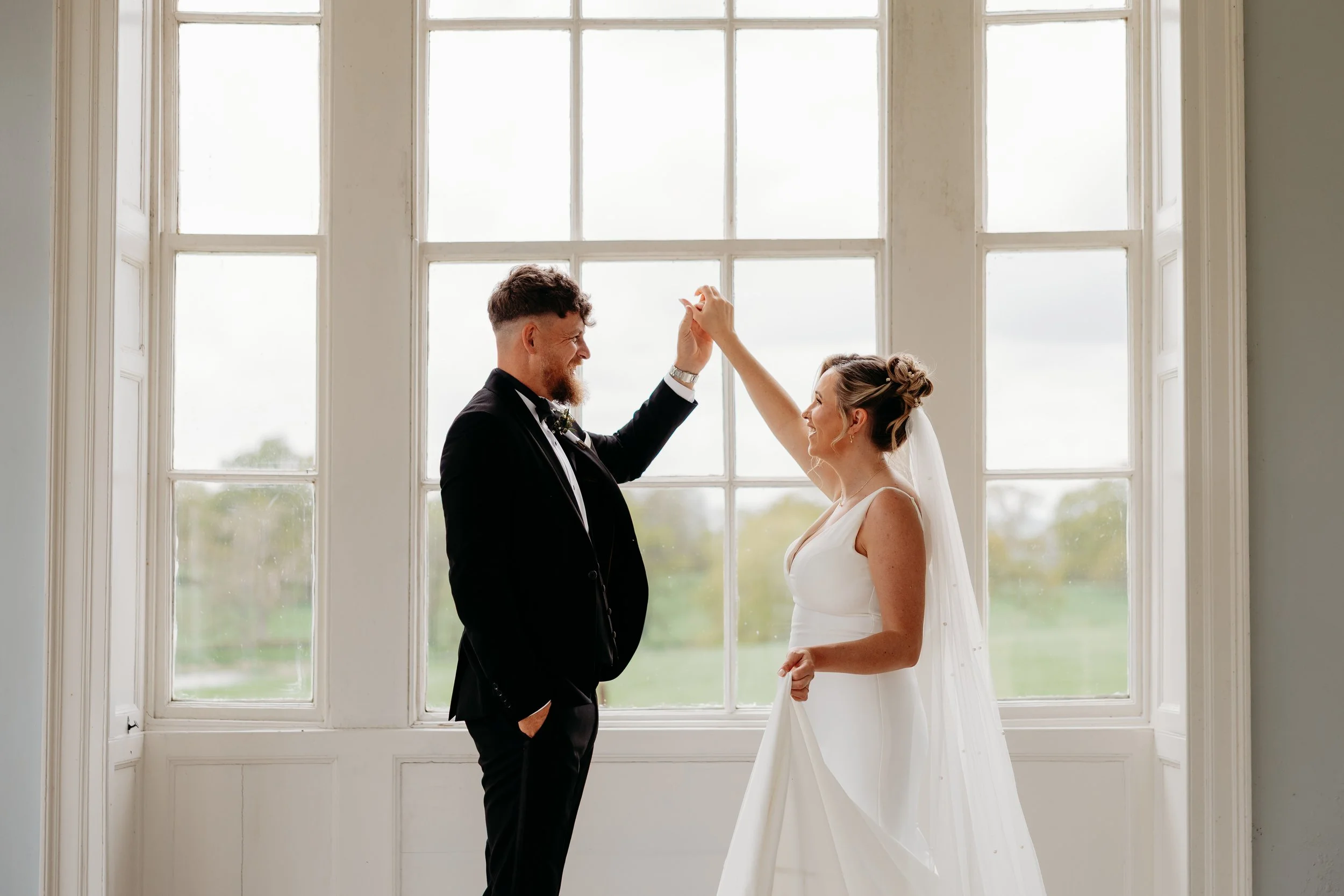 A bride and groom dancing together in front of a large window, with the groom in a black tuxedo and the bride in a white wedding dress, during their wedding celebration.