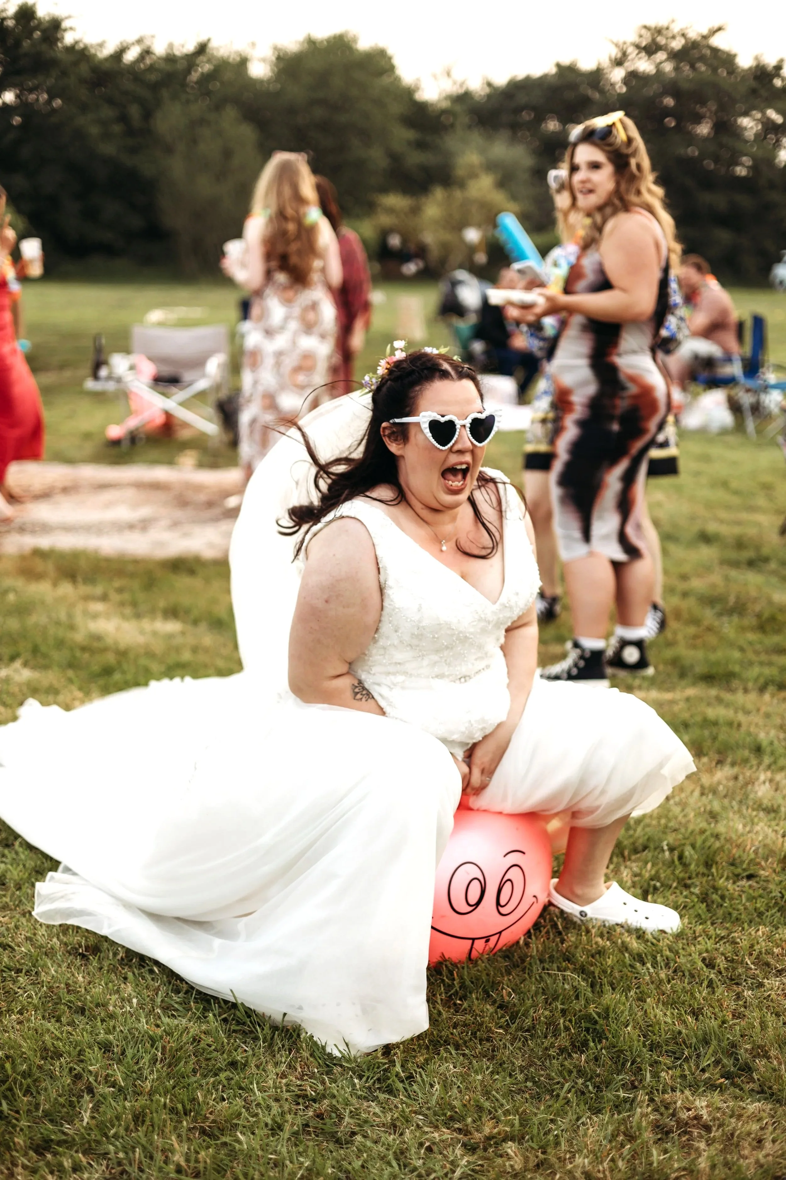 A woman in a wedding dress and heart-shaped sunglasses humorously sits on a pink pig-shaped balloon during an outdoor celebration, with other people in the background.