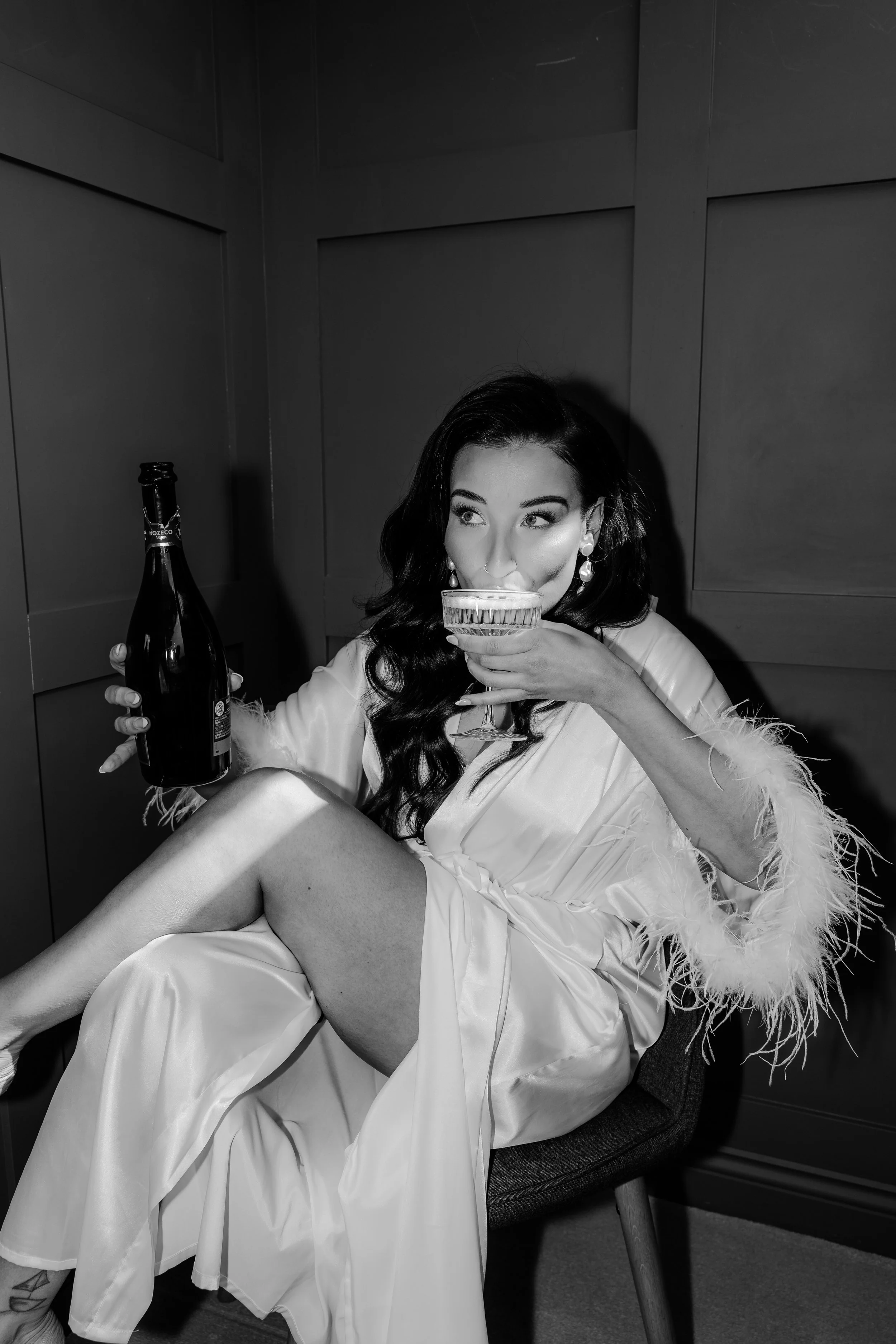 Black and white photo of a woman sitting on a chair, wearing a satin robe with feather trim, holding a champagne bottle in her left hand and a cocktail glass in her right hand, near her lips.