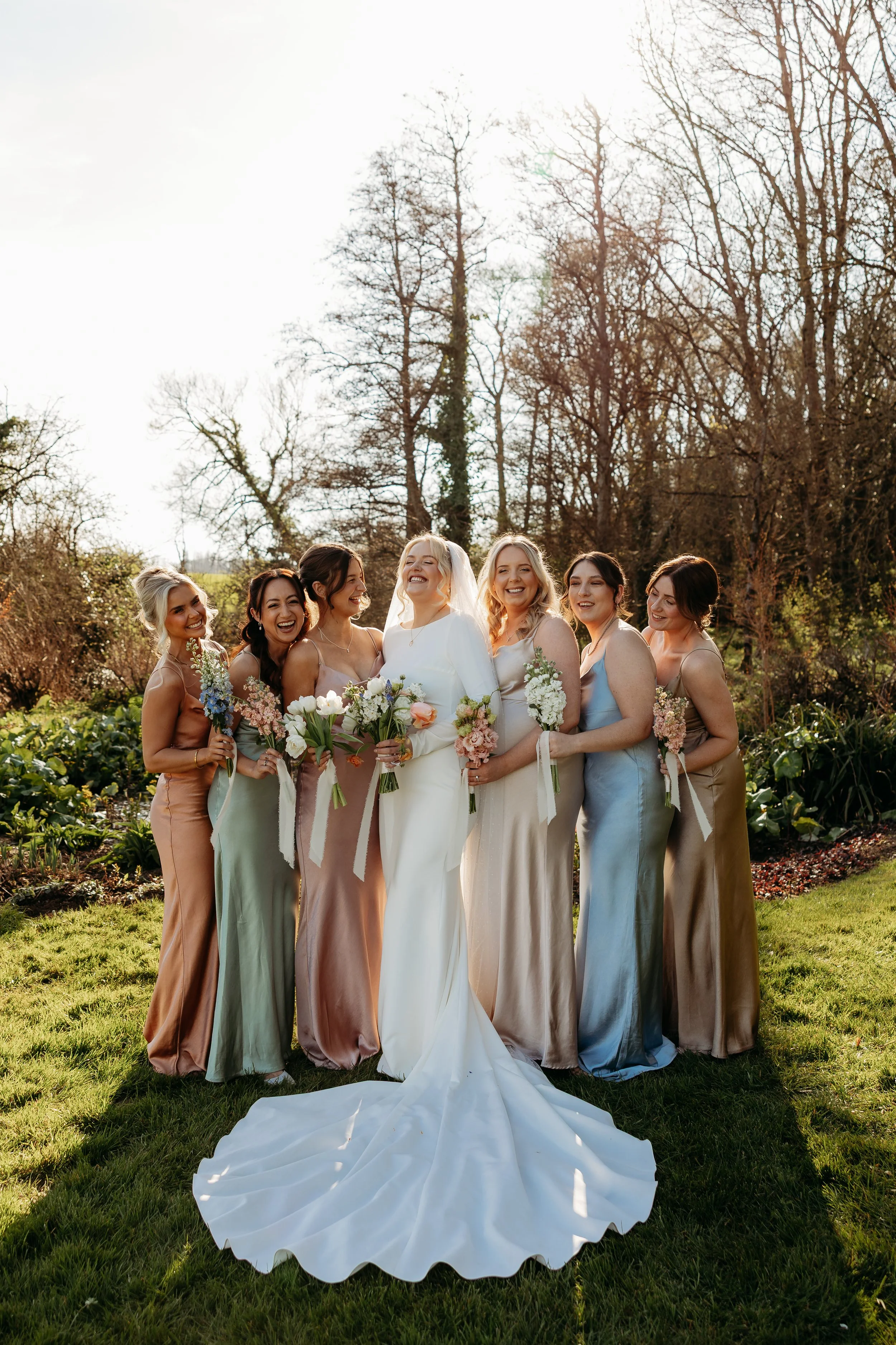 A bride in a white wedding dress and veil stands in the center of a group of six women dressed in pastel satin dresses, outdoors on a sunny day with a garden background, all smiling and holding bouquets of flowers.