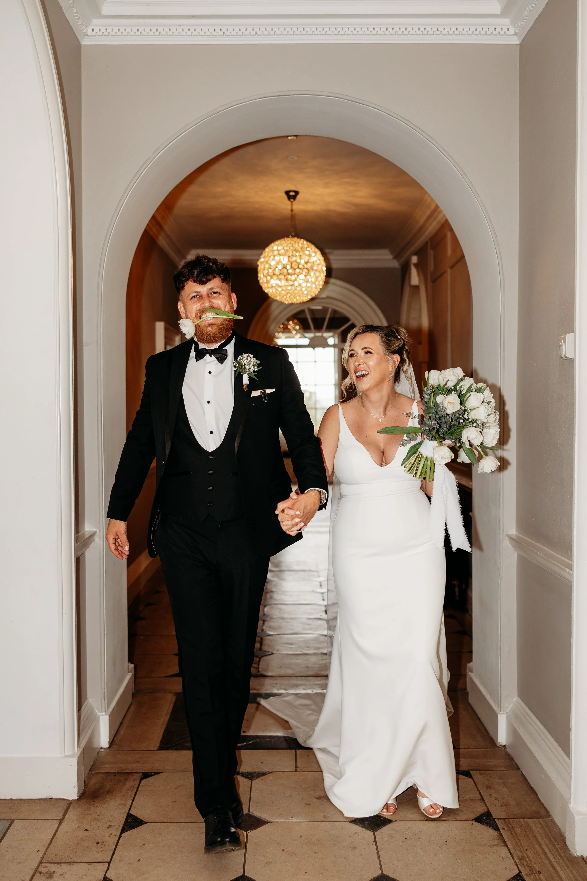 A newlywed couple walking through an arched hallway. The groom has a beard, is smiling, and holds a white flower in his mouth. He is wearing a tuxedo. The bride is laughing, holding a bouquet of white and green flowers, and wearing a white wedding go