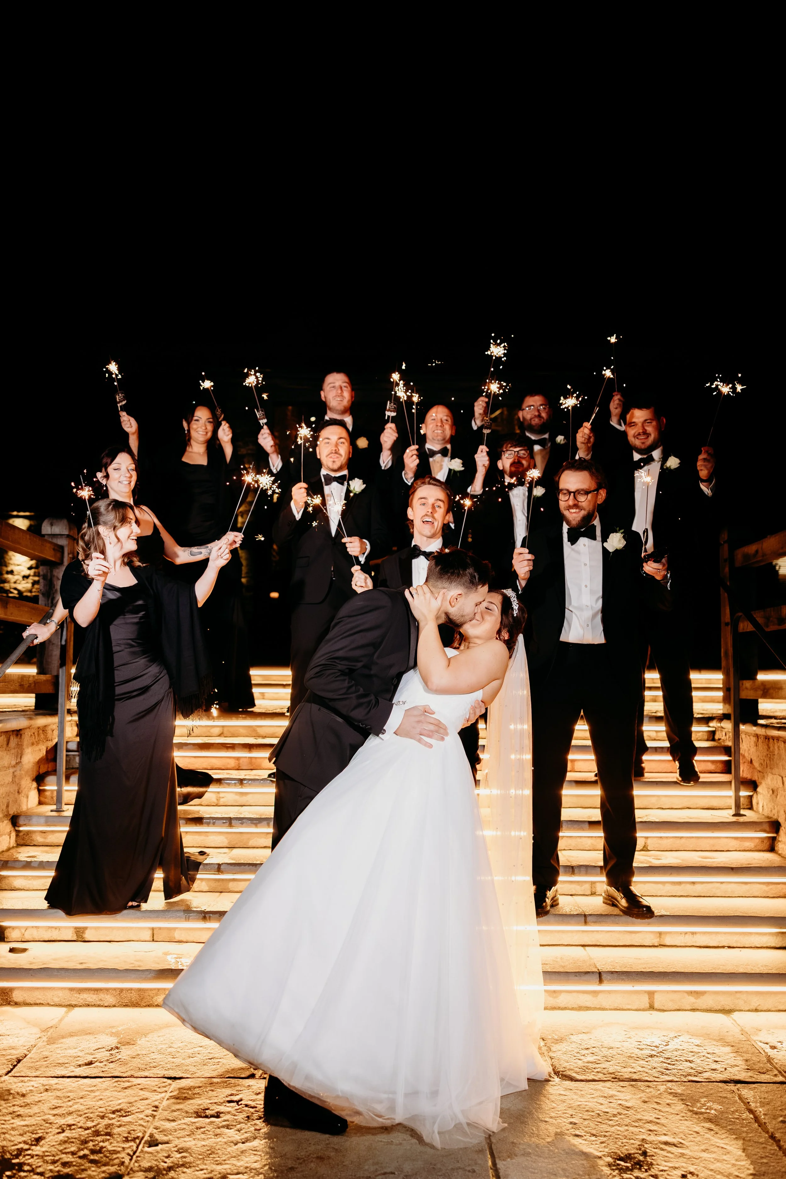 A bride and groom share a kiss in front of their wedding party on illuminated stairs, with the wedding party holding sparklers at night. Creating beautiful moments in the Cotswolds