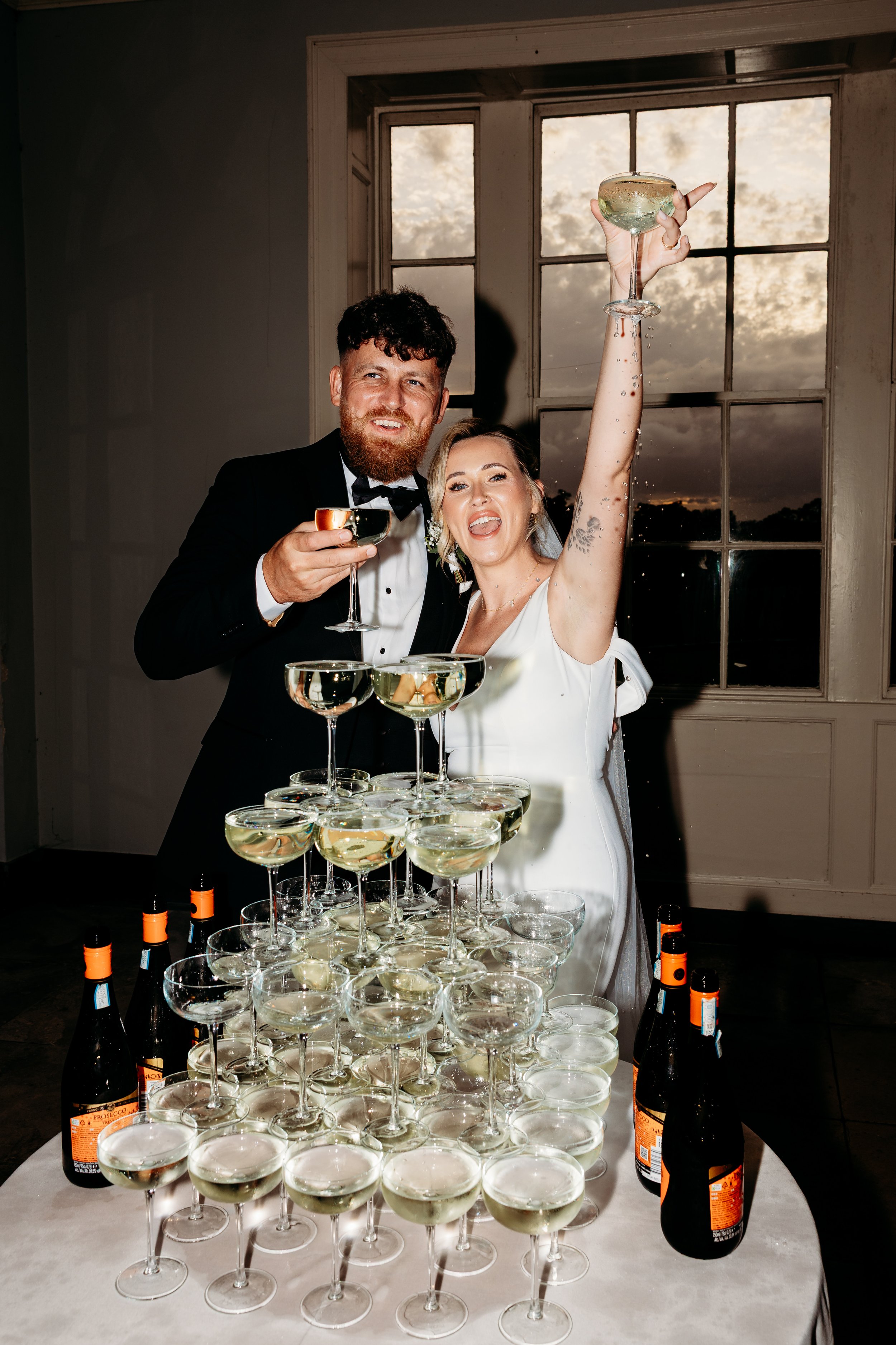 A couple dressed in wedding attire celebrating with champagne, raising glasses, and standing behind a pyramid of champagne glasses and bottles on a table.