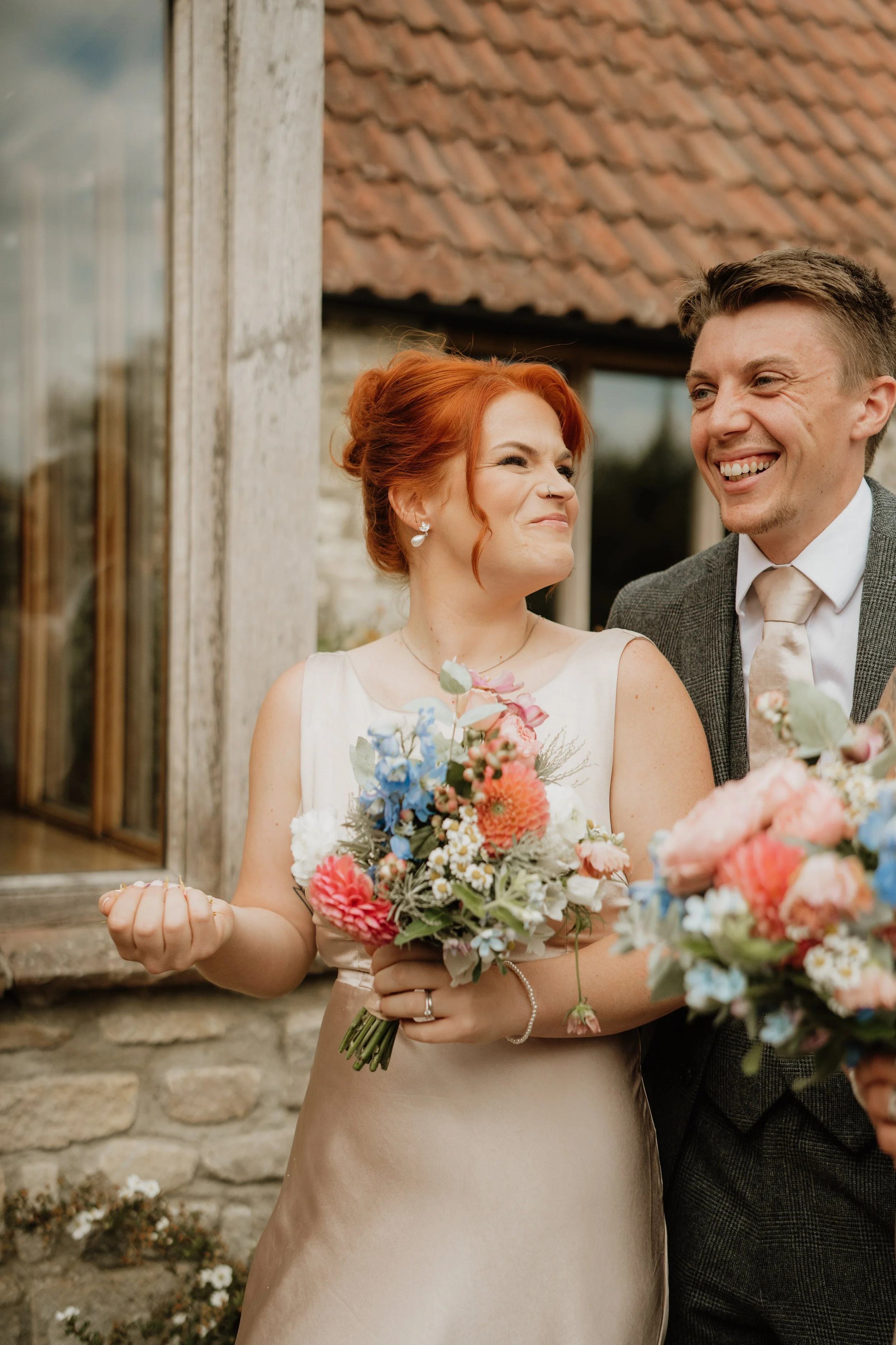 A woman with red hair wearing a cream-colored dress holding a colorful bouquet of flowers, smiling beside a man in a gray suit with a tie, standing outside a rustic building with a stone foundation and wooden window.
