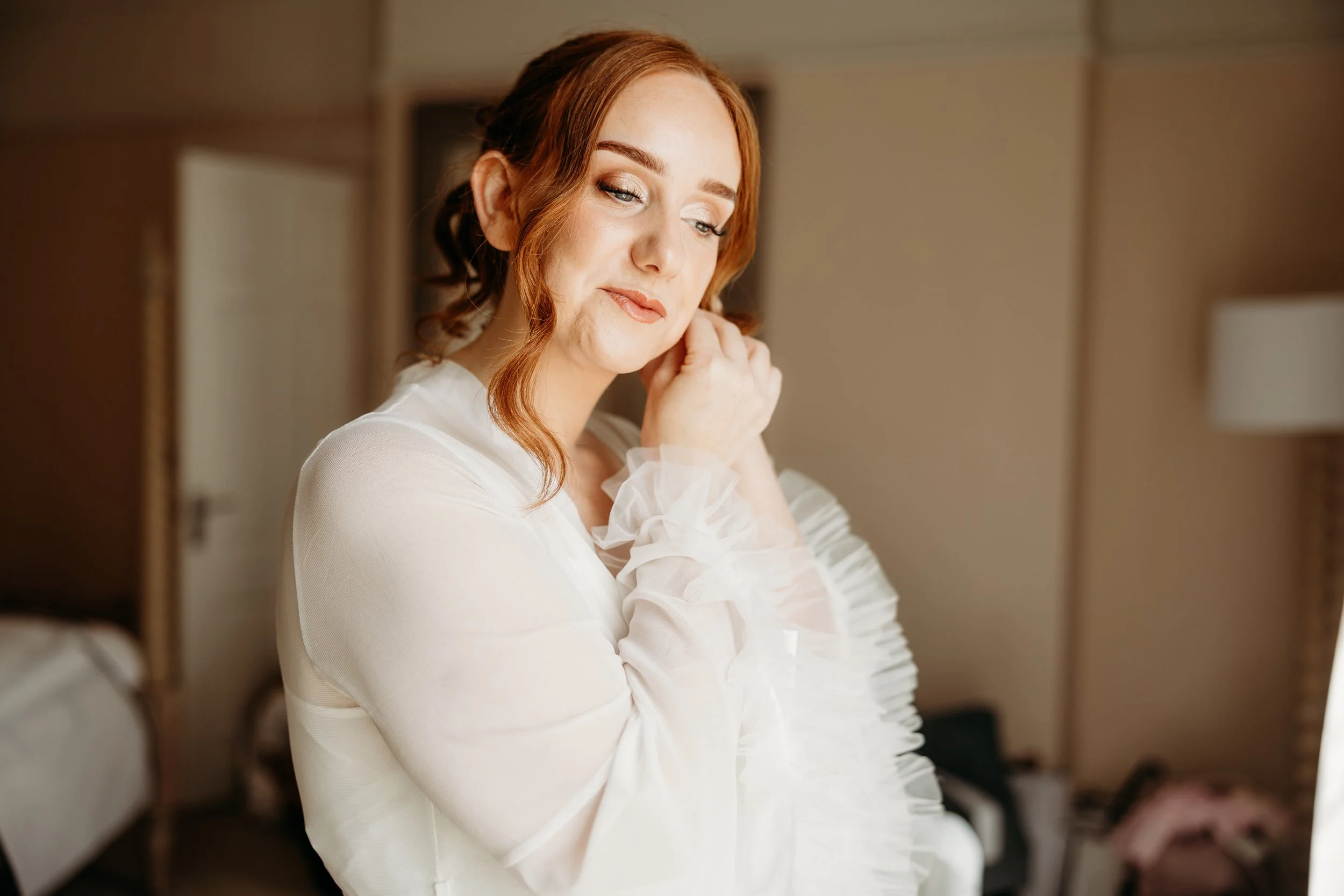 A woman with red hair and makeup adjusts her earring in a softly lit room, wearing a sheer white top with ruffled sleeves.