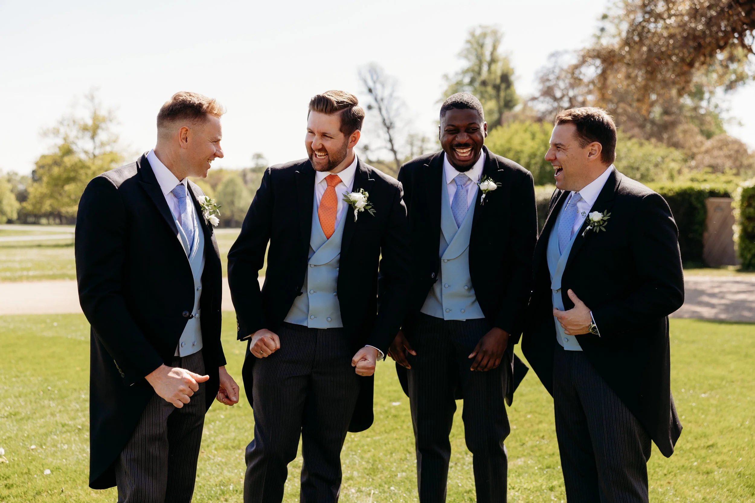 Four men in formal suits standing outside on a sunny day, smiling and interacting with each other at Goodwood Estate, Sussex.