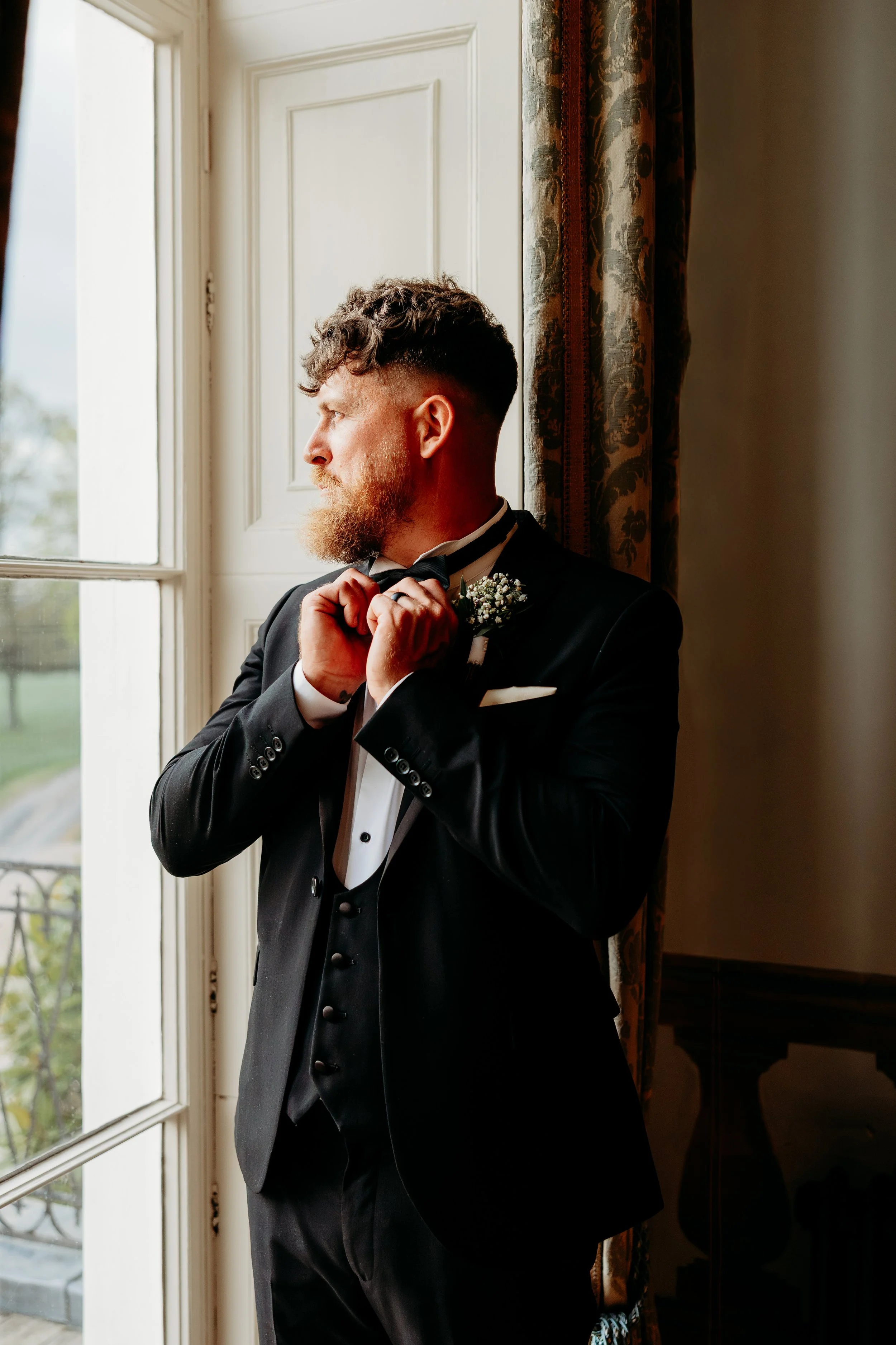 Man with curly hair and a beard adjusting his bowtie while standing by a large window with floral curtains.