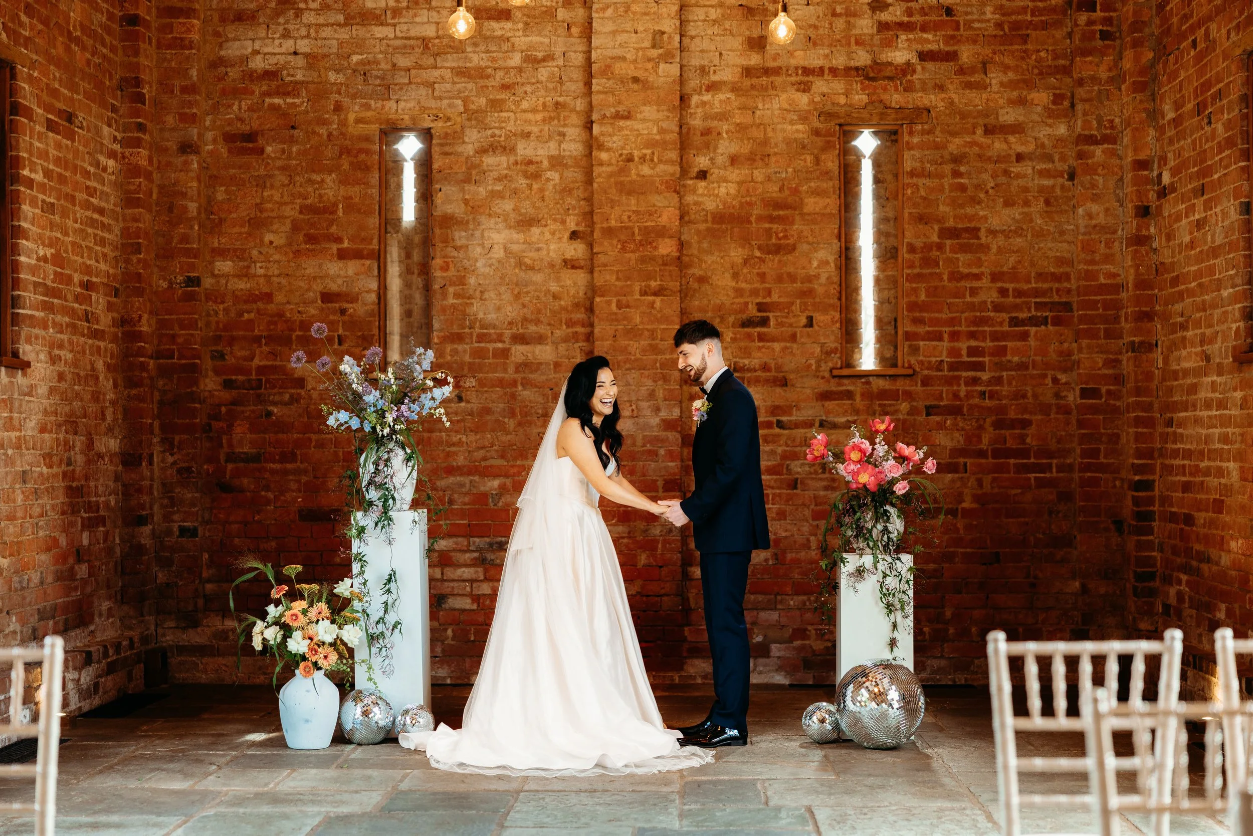 A bride and groom holding hands during their wedding ceremony inside a brick-walled venue, decorated with floral arrangements and disco balls.