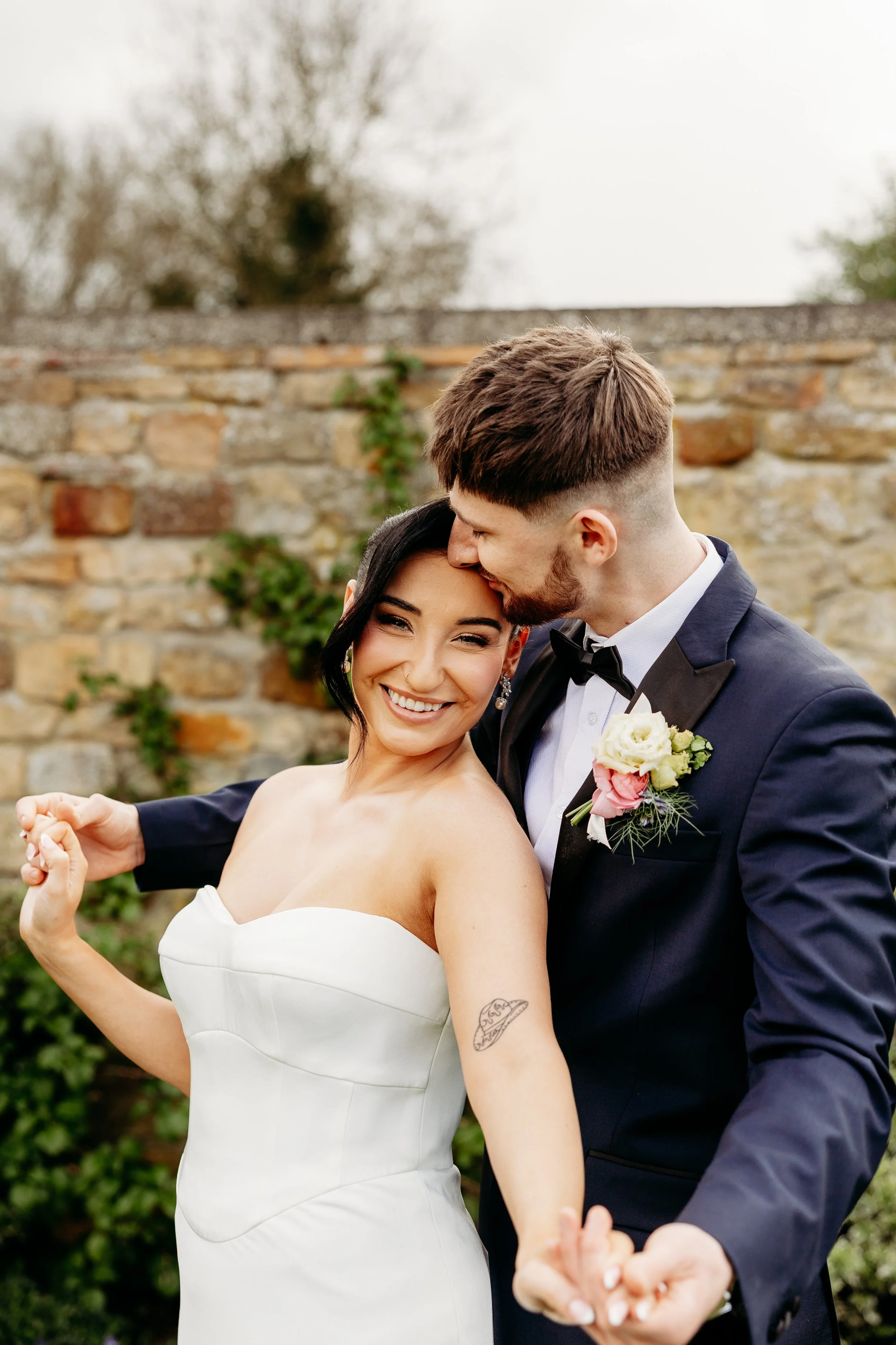 A happy bride and groom dancing outdoors, with the groom kissing the bride on the forehead and the bride smiling joyfully. The bride is in a strapless white wedding dress, and the groom is in a navy tuxedo with a boutonniere. They are standing in fro