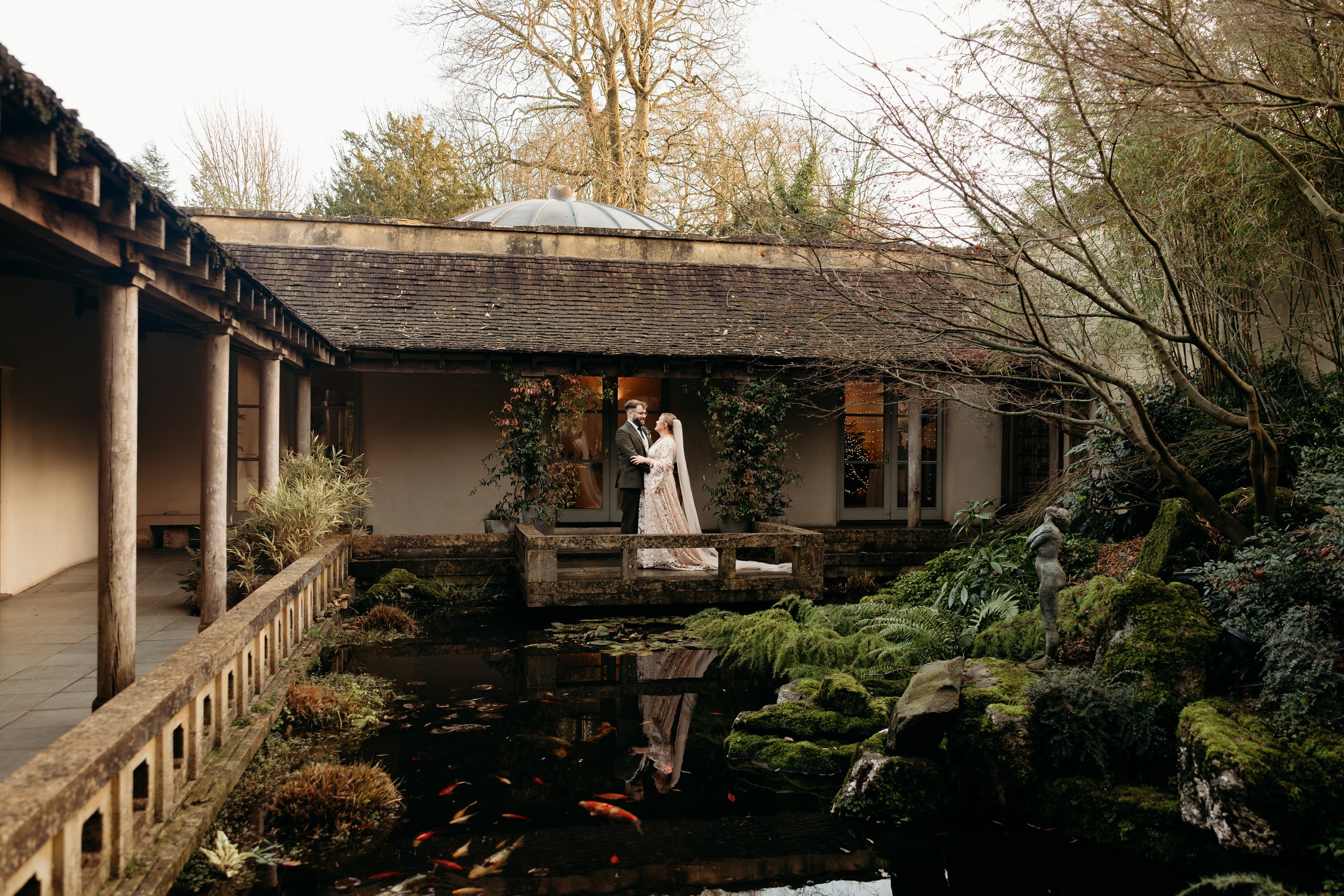 A bride and groom standing on a small stone bridge over a pond, surrounded by a garden with moss-covered rocks, plants, and a statue, in front of a rustic building with large glass doors and trees in the background.
