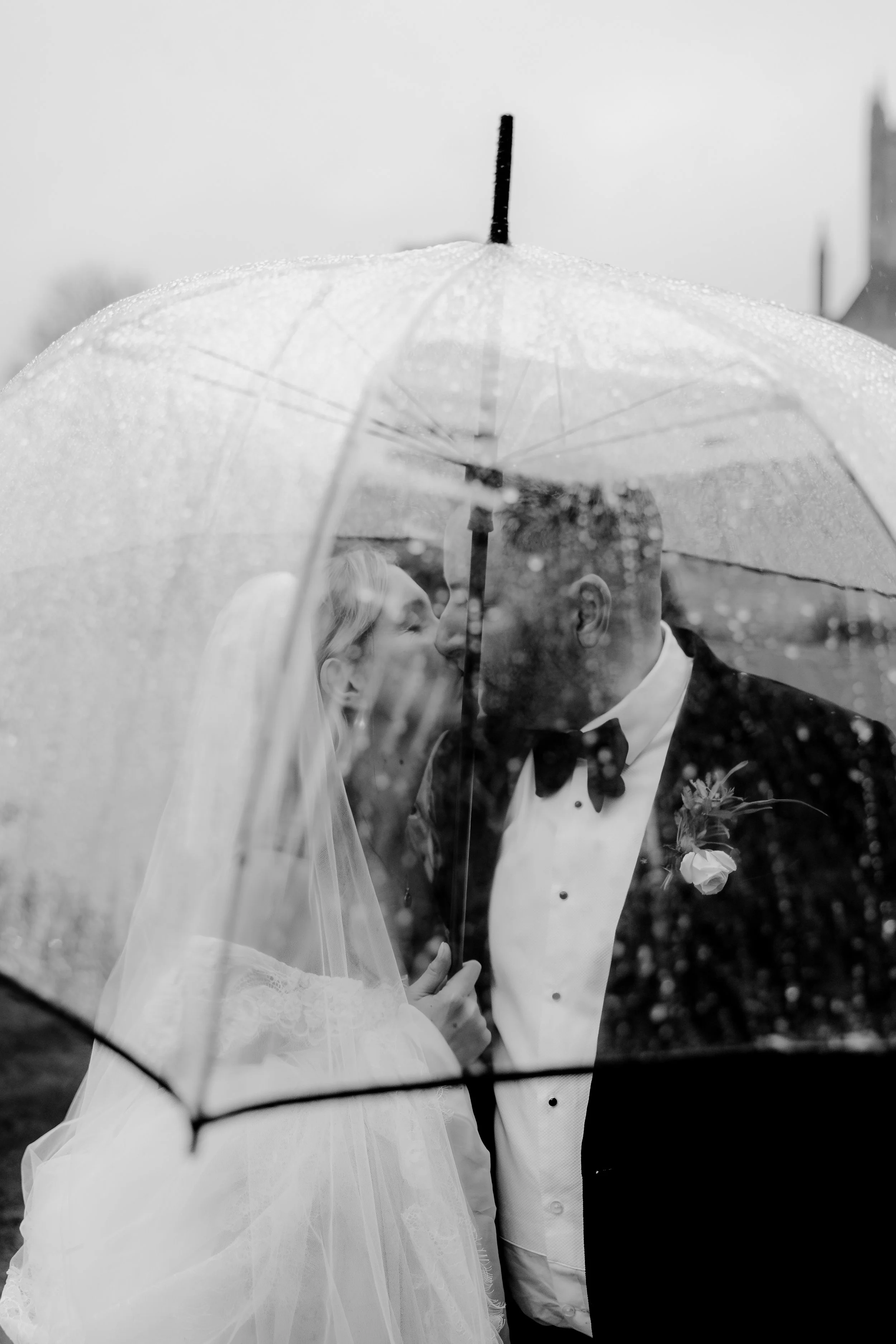 A black-and-white photo of a bride and groom sharing a kiss under a transparent umbrella on a rainy day.
