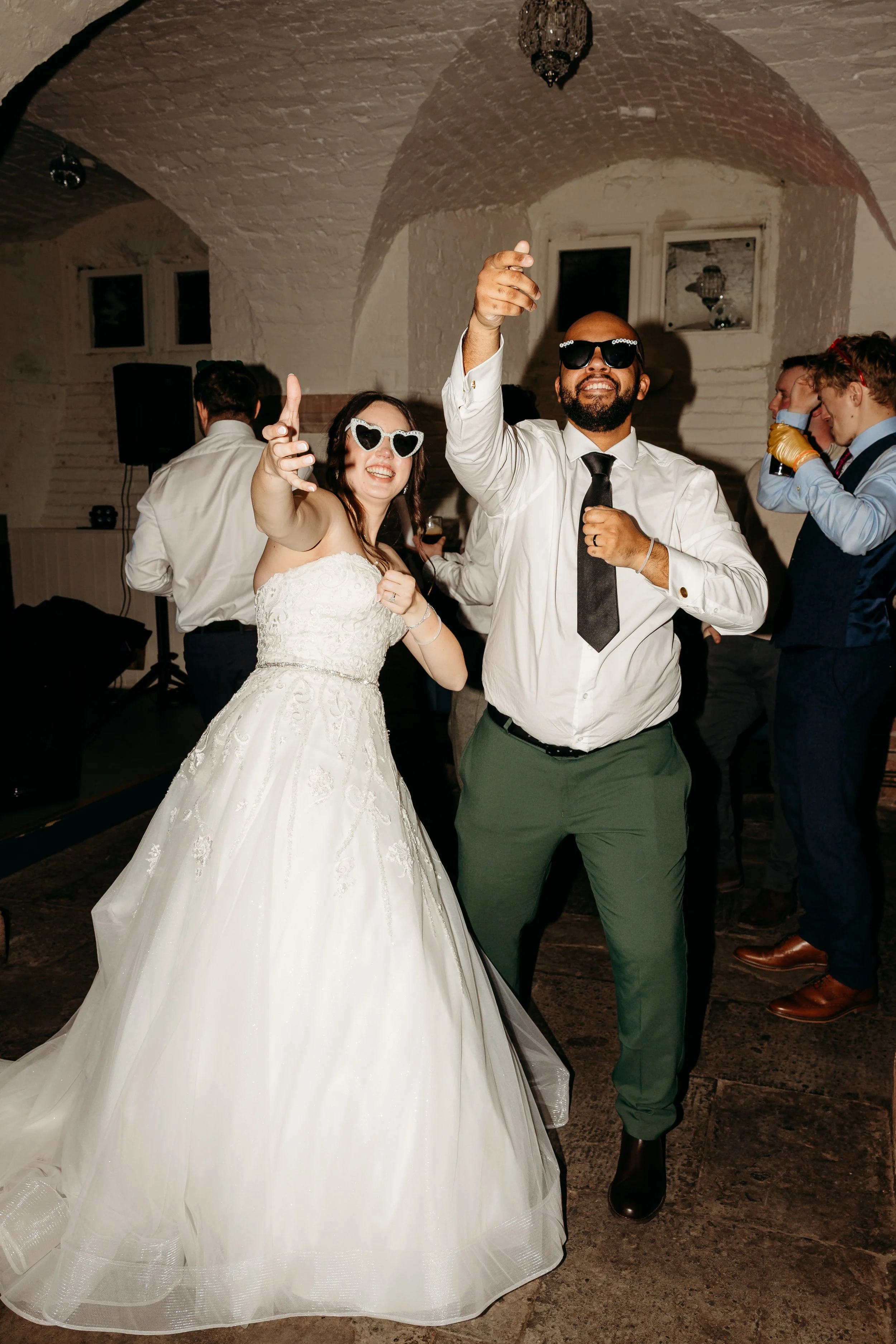 People dancing and celebrating at a wedding reception in a rustic venue with white brick walls. A bride in a wedding dress and a man in a white shirt with a black tie are wearing sunglasses and smiling.
