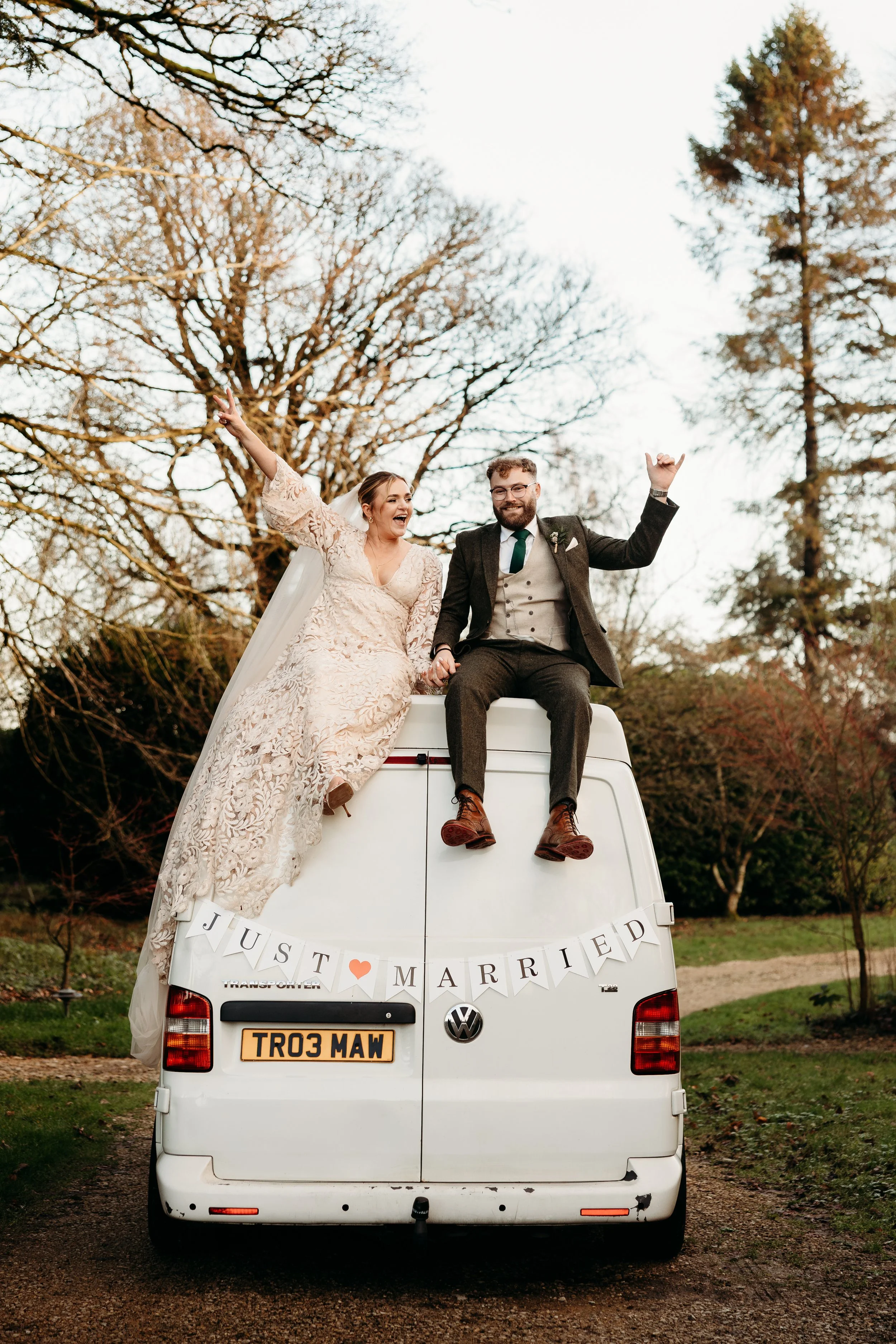A newly married couple sitting on top of a white van, celebrating their wedding outdoors. The bride is wearing a lace wedding dress and the groom is in a suit. A 'Just Married' banner is displayed on the back of the van, and the couple is smiling and