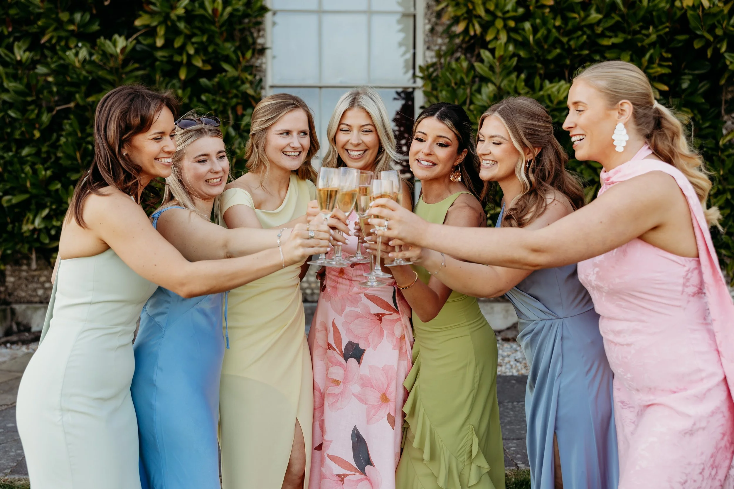 Eight women in colorful dresses holding glasses of champagne in a toast, standing outdoors near a green hedge at Goodwood Estate, Sussex.