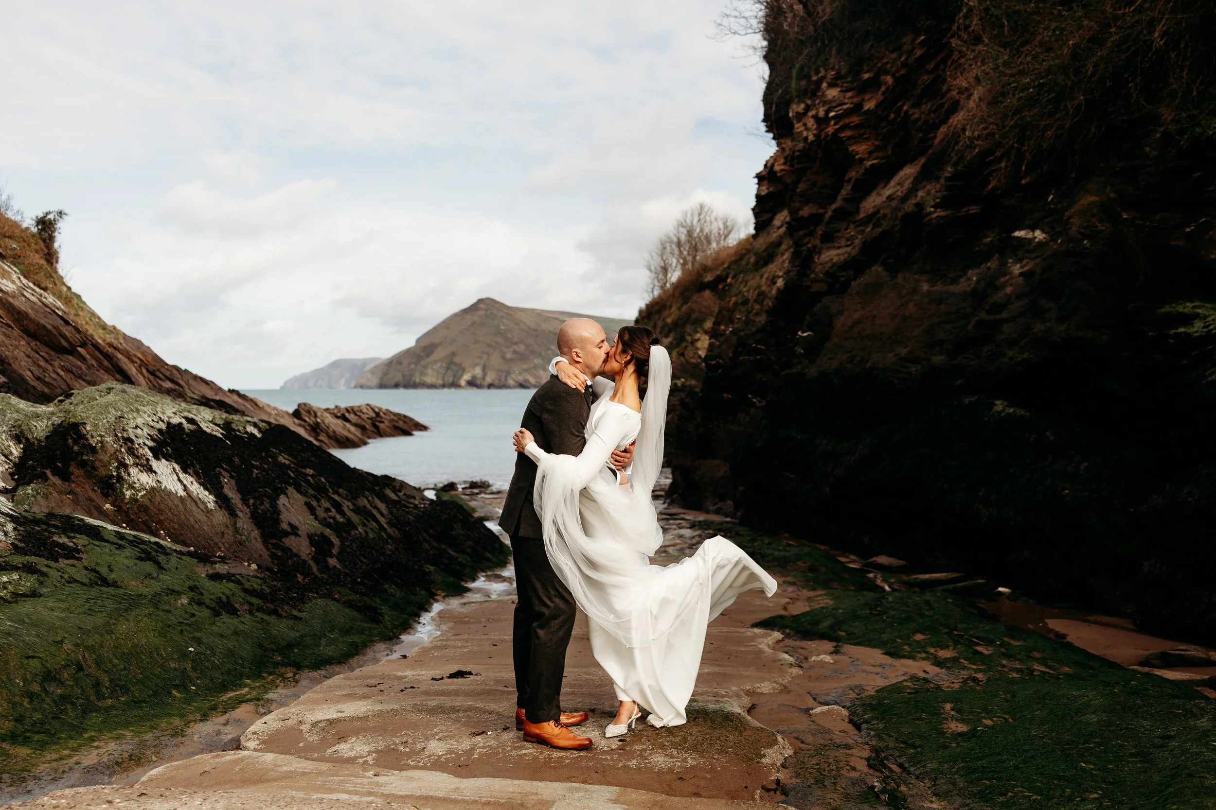 A couple dressed in wedding attire sharing a kiss on a rocky beach with cliffs and the ocean in the background.