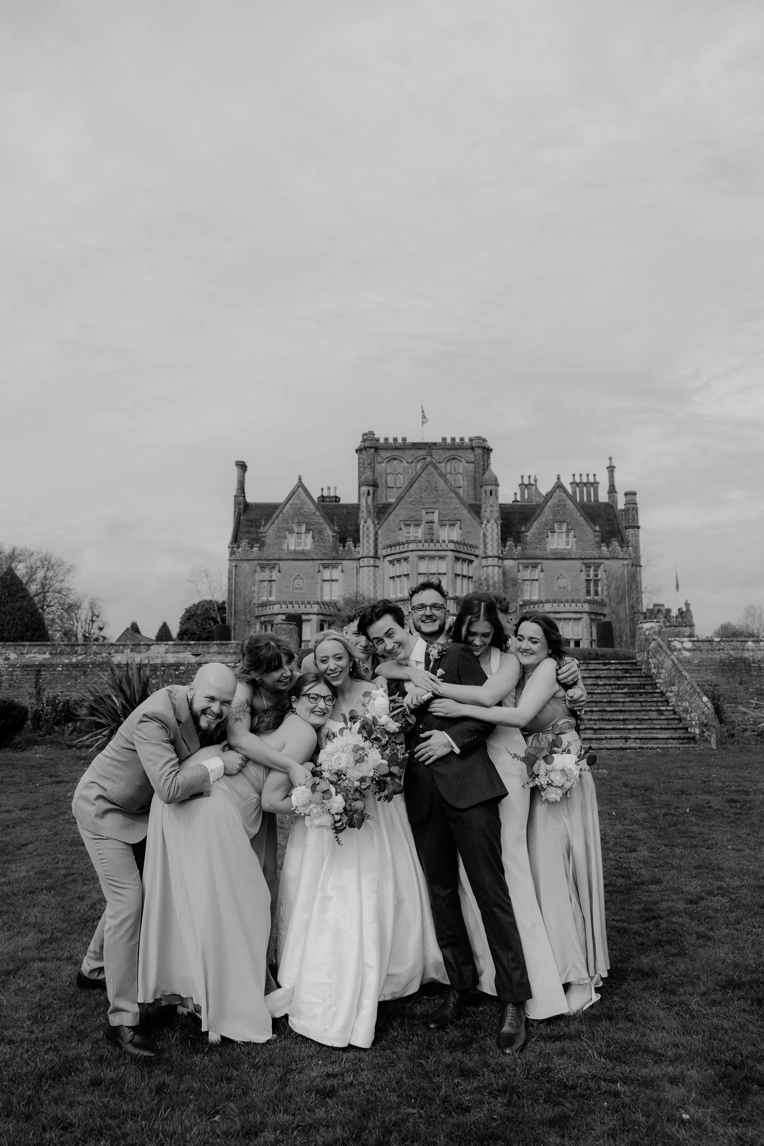 A group of wedding guests, including bridesmaids and groomsmen, happily hugging and celebrating on the lawn of a historic castle-like building.