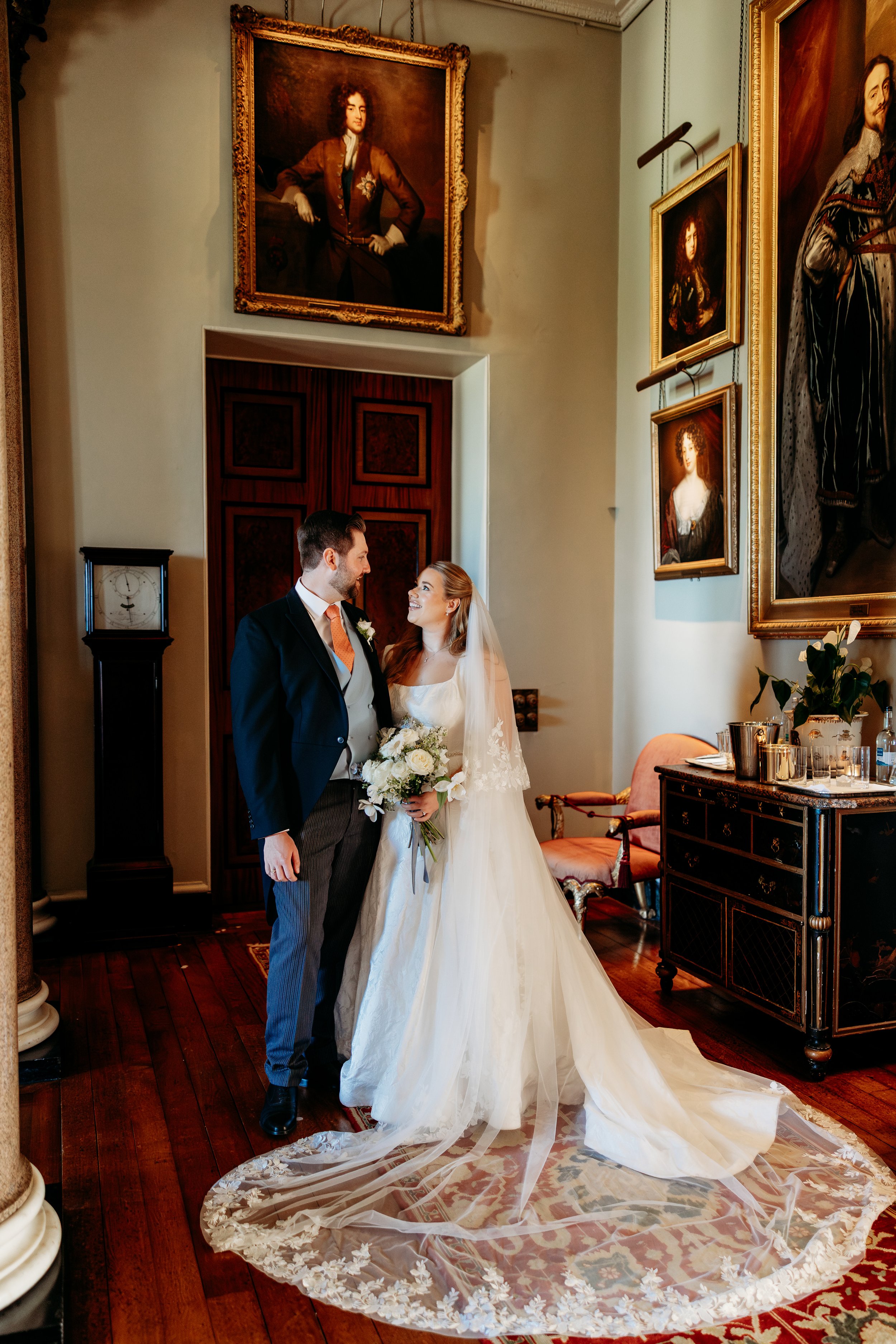 A bride and groom standing inside an elegant room with large portraits on the wall. The bride is wearing a white wedding dress with a long veil and holding a bouquet of white flowers at Goodwood Estate, Sussex.