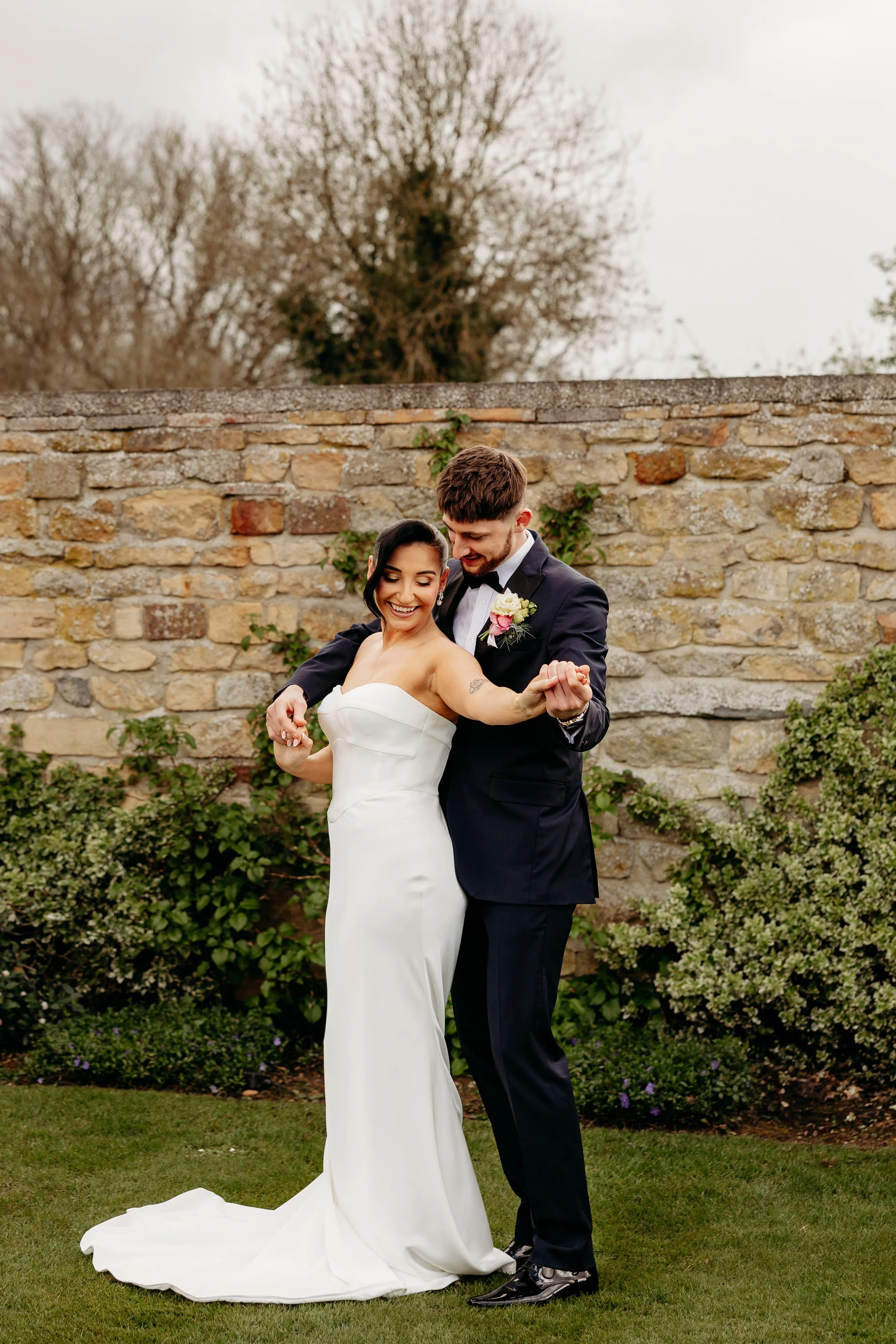 A newlywed couple dancing outdoors in front of a stone wall, with trees in the background. The bride is in a white strapless wedding gown and the groom is in a black tuxedo with a boutonniere.